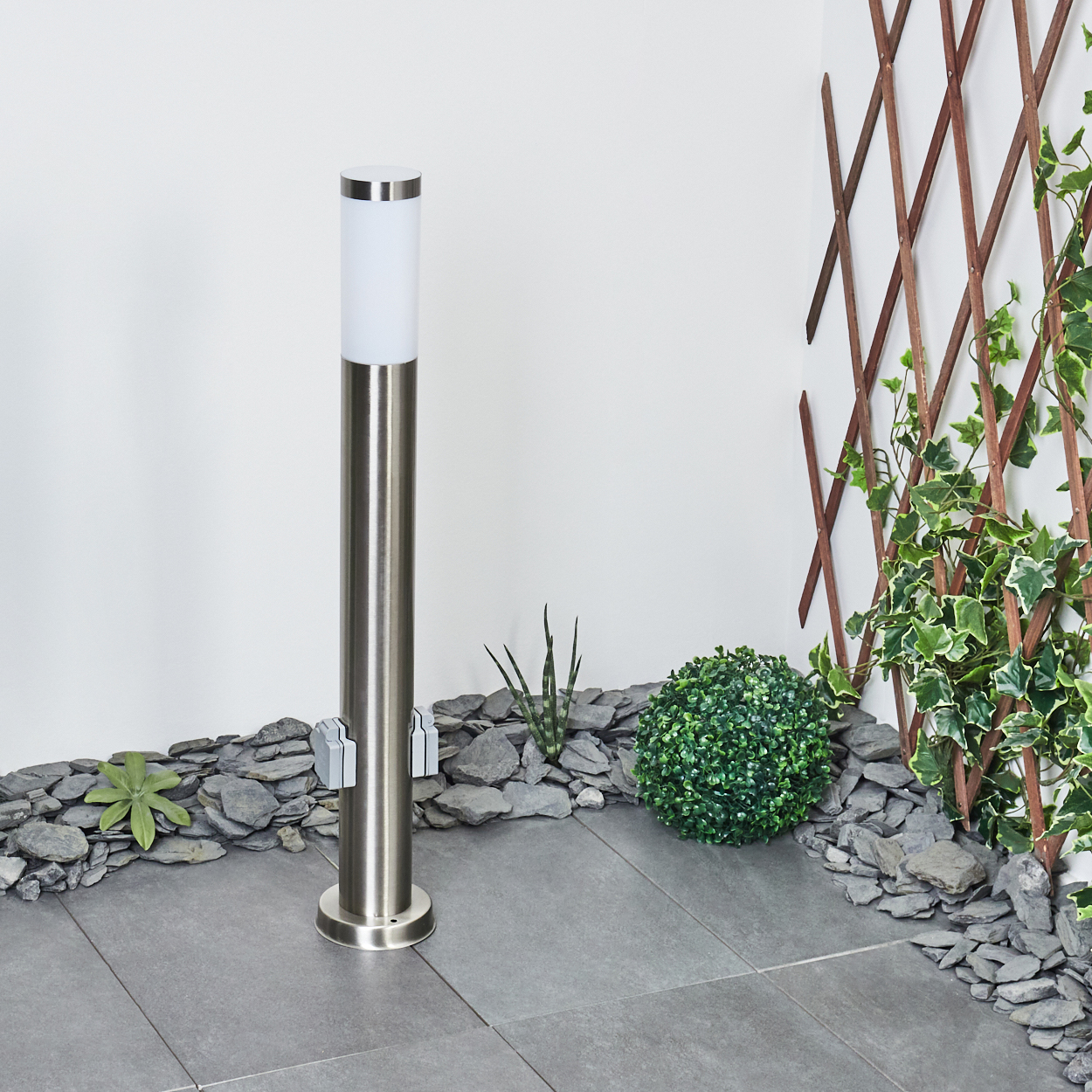 Eine moderne Außenleuchte steht in einem Garten. Sie ist von Steinen, Pflanzen und einem Holzgitter mit Efeu umgeben.