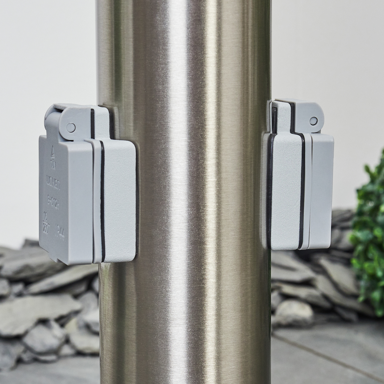 Eine Edelstahlstange mit zwei grauen Elektrobox-Aufsätzen. Der Hintergrund umfasst Schiefer und grünes Laub.