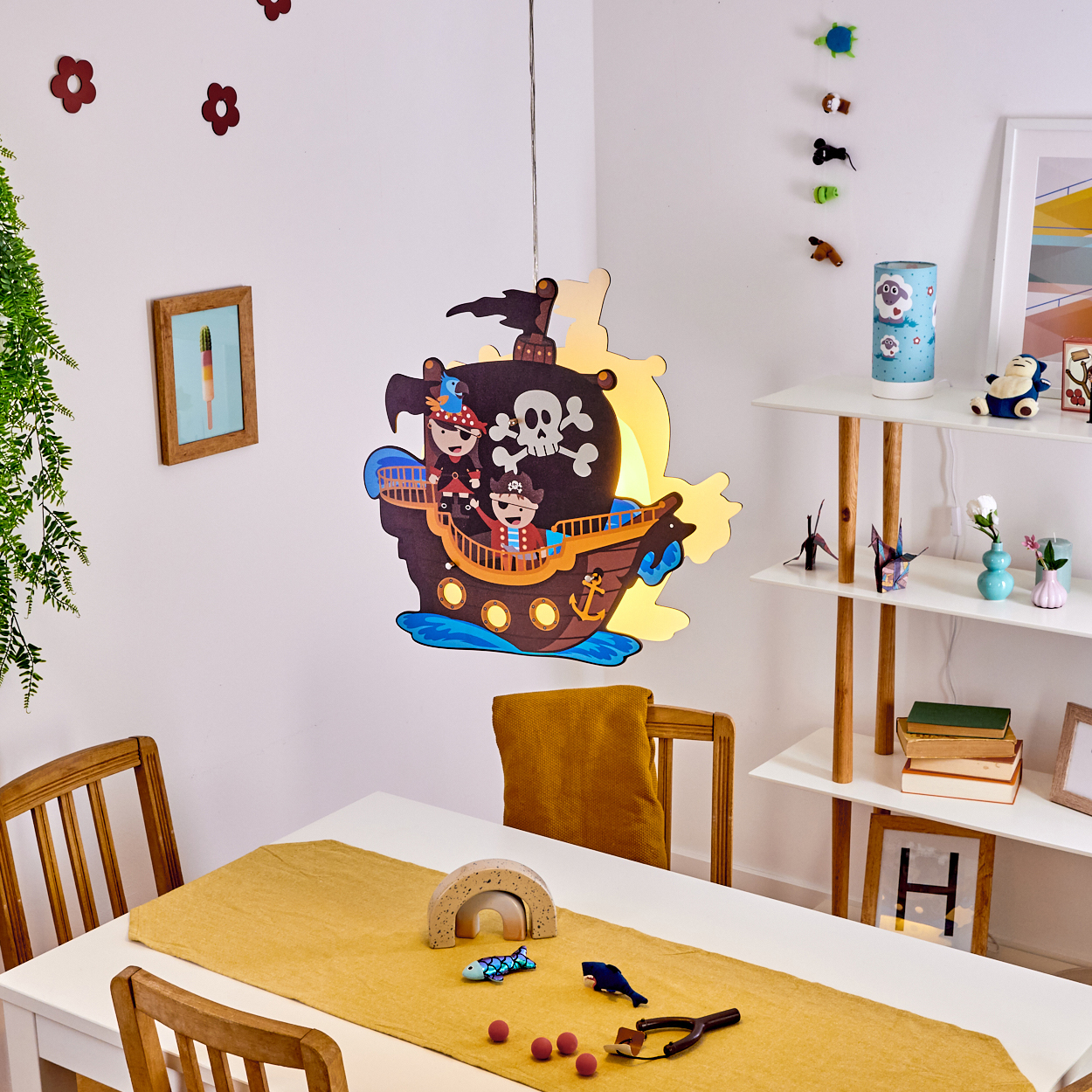 Über einem Tisch mit Spielzeug hängt eine Piratenschifflampe. Ein Regal beherbergt Bücher und Dekorationen. Holzstühle umgeben den Tisch.