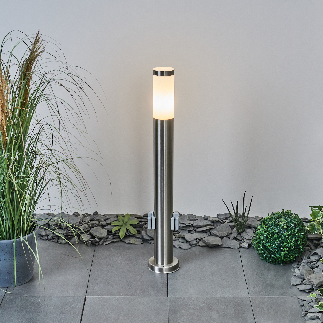 Eine beleuchtete, silberne Außenlampe steht auf grauen Fliesen mit dekorativen Steinen und Pflanzen. Hohes Gras steht links in einem Topf.