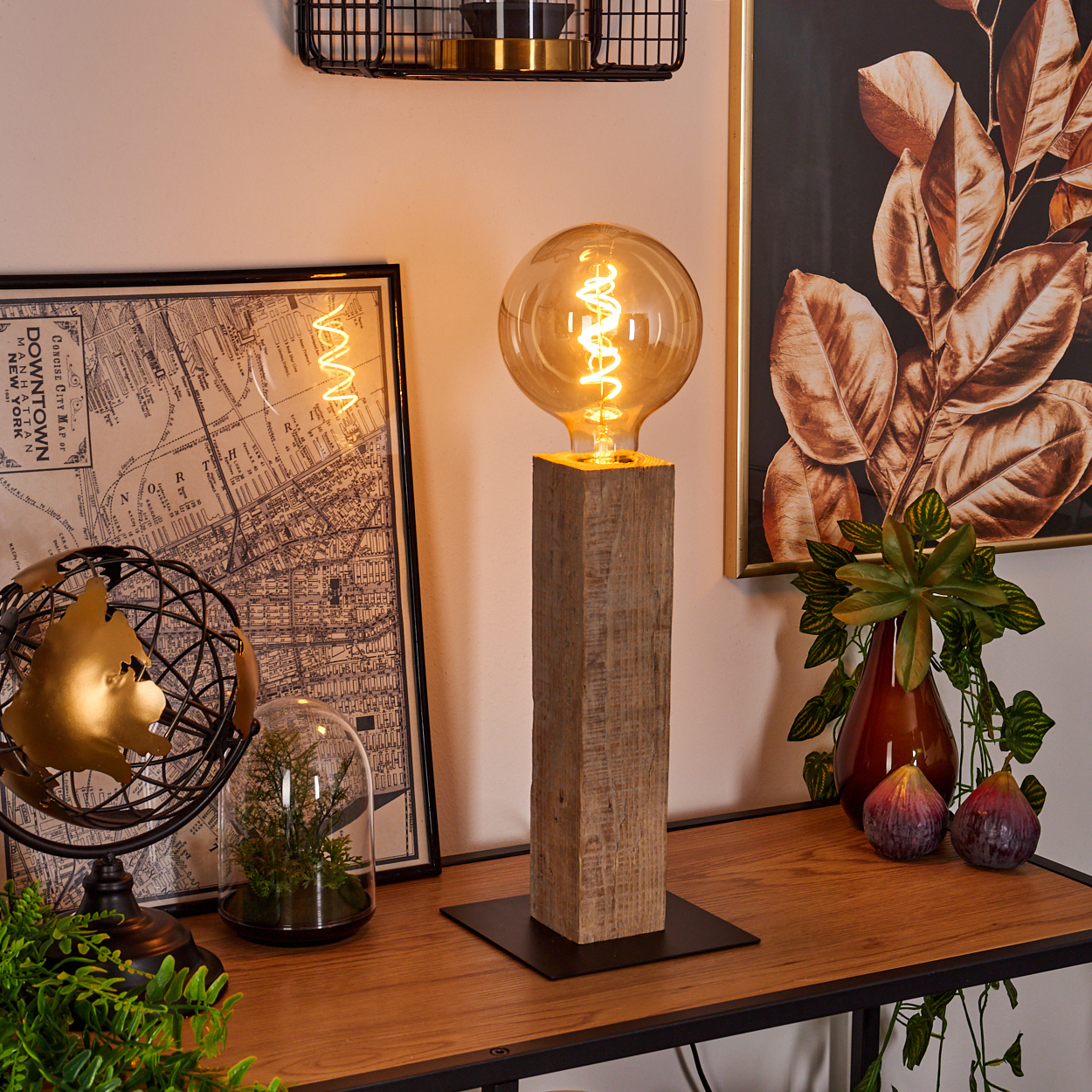 Eine beleuchtete Glühbirnenlampe auf einem Holzsockel, Karte, Globus, Pflanzen und gerahmte Kunst auf einem Holztisch. Der Hintergrund ist beige.