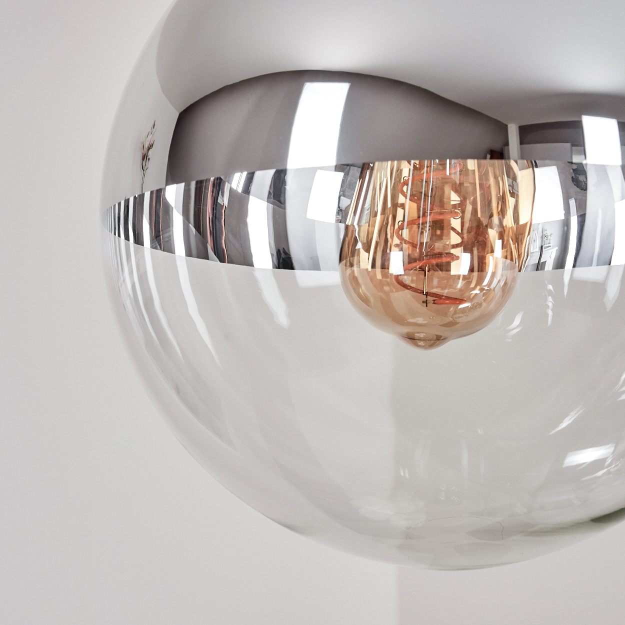 Nahaufnahme einer modernen Leuchte. Eine klare Glaskugel mit Chromband, die eine warmtonige Glühbirne enthält, vor weißem Hintergrund.