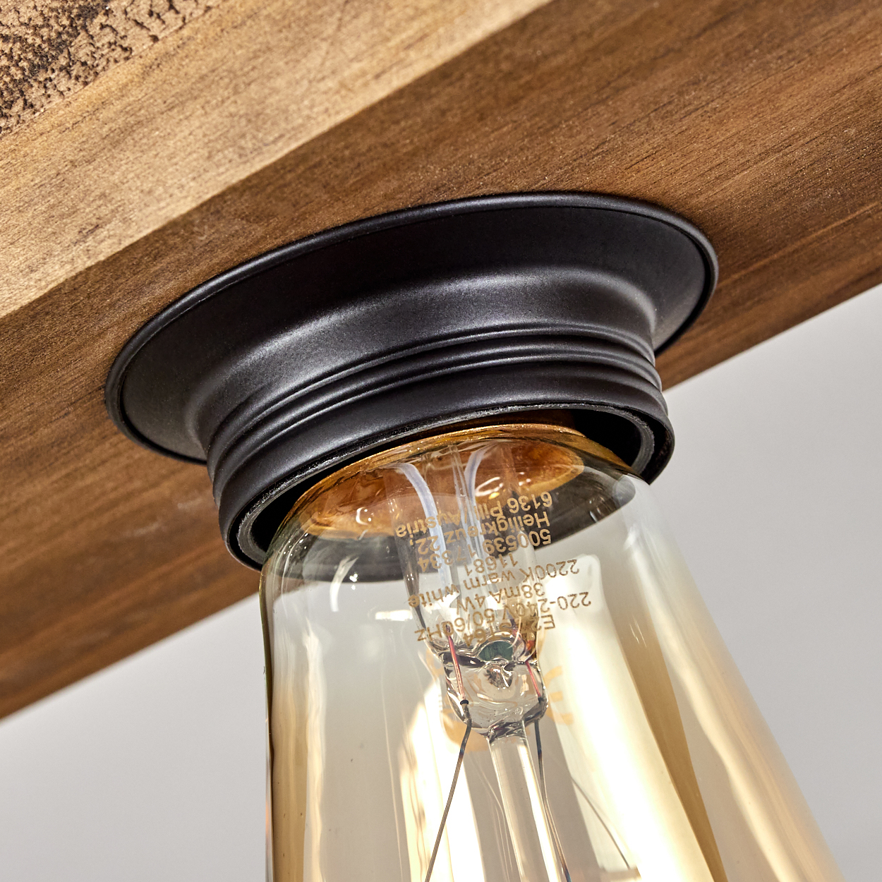 Nahaufnahme einer Leuchte mit einer Glühbirne im Vintage-Stil. Sie ist an einem Holzbalken mit schwarzem Metallsockel befestigt. Der Hintergrund ist weiß.