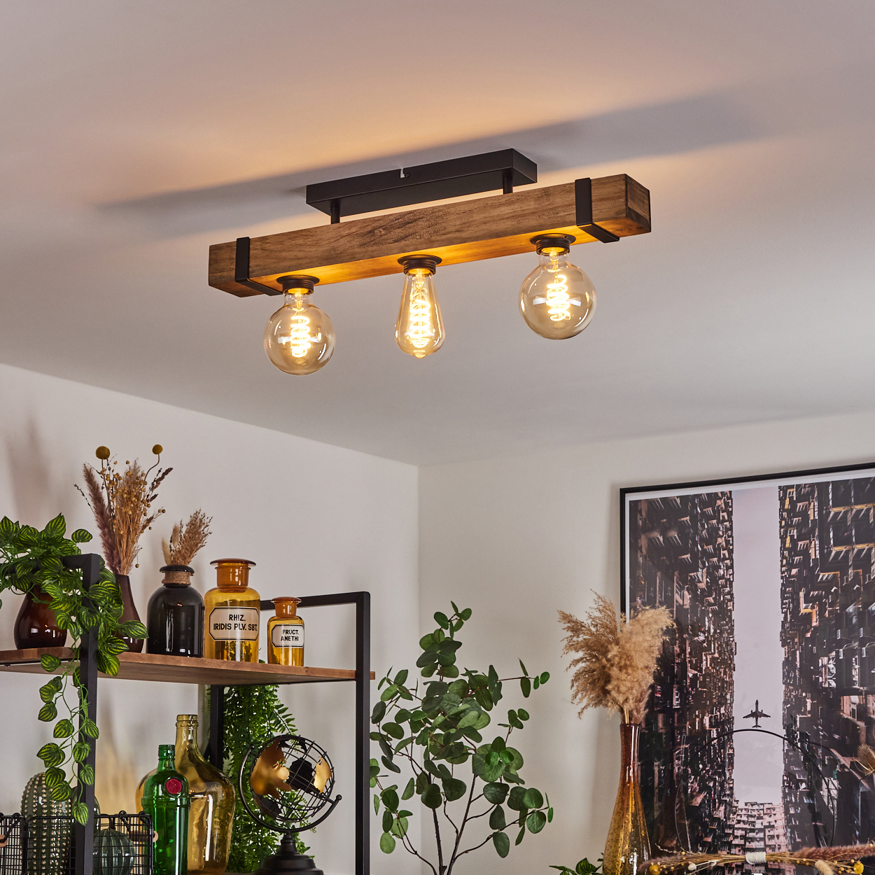 Eine Deckenlampe aus Holzbalken mit drei leuchtenden Glühbirnen. Darunter Pflanzen und Dekorationsartikel. Ein Stadtlandschaftskunstwerk hängt an der Wand.