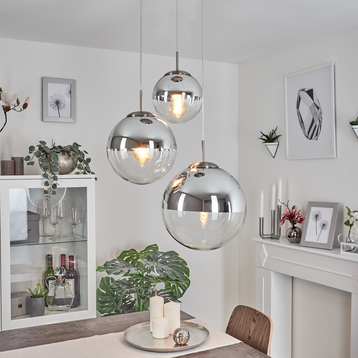 Drei Pendelleuchten aus Chrom und Glas hängen über einem Tisch. Pflanzen und gerahmte Kunst schmücken den Raum mit weißen Wänden.