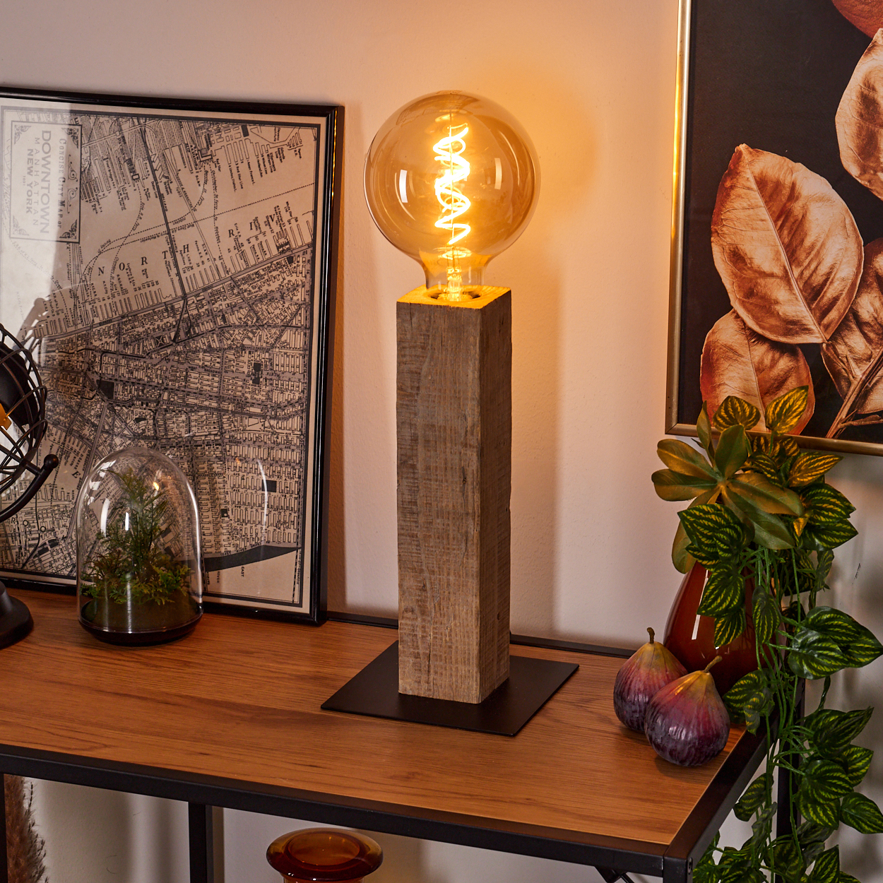 Eine Lampe mit leuchtender Glühbirne steht auf einem Holzblock. Eine Karte, Kunst, Pflanzen und Feigen stehen auf einem Holztisch. Der Hintergrund ist eine Wand.