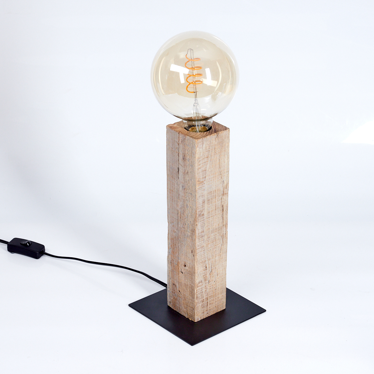 Eine Holzlampe mit einer großen Glühbirne. Sie steht auf einem schwarzen quadratischen Sockel, mit Netzkabel und Schalter. Der Hintergrund ist weiß.