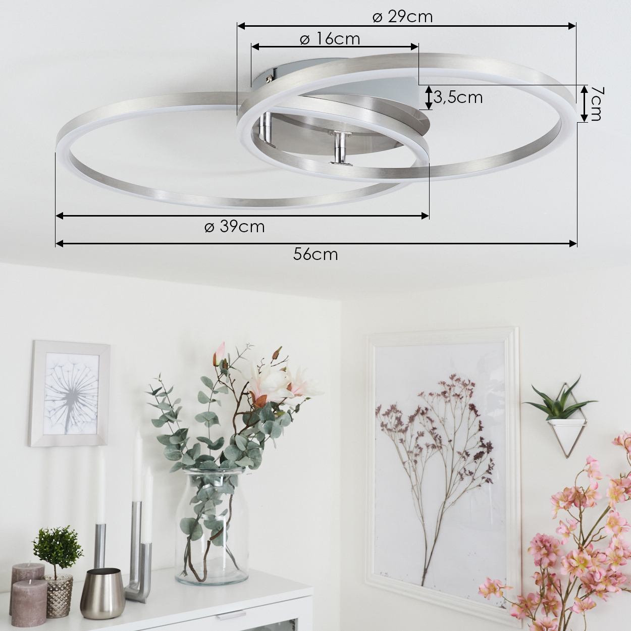 Eine moderne Deckenleuchte mit drei silbernen Ringen. Maße sind angegeben. Darunter eine weiße Wand mit Bildern, Blumen und Kerzen.
