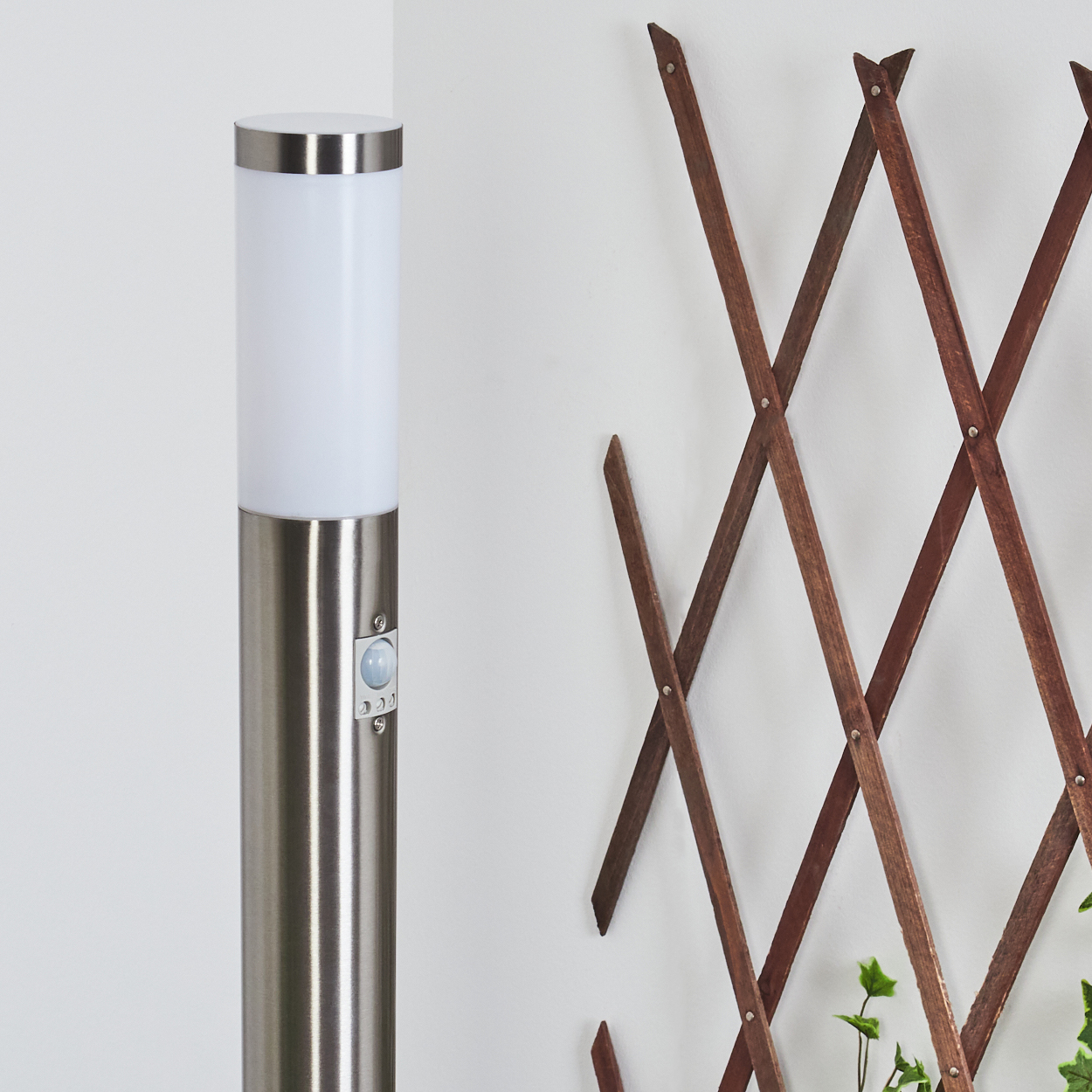 Eine metallische Außenlampe und ein Holzgitter vor einer weißen Wand. Die Lampe hat einen weißen Deckel und einen Bewegungssensor.