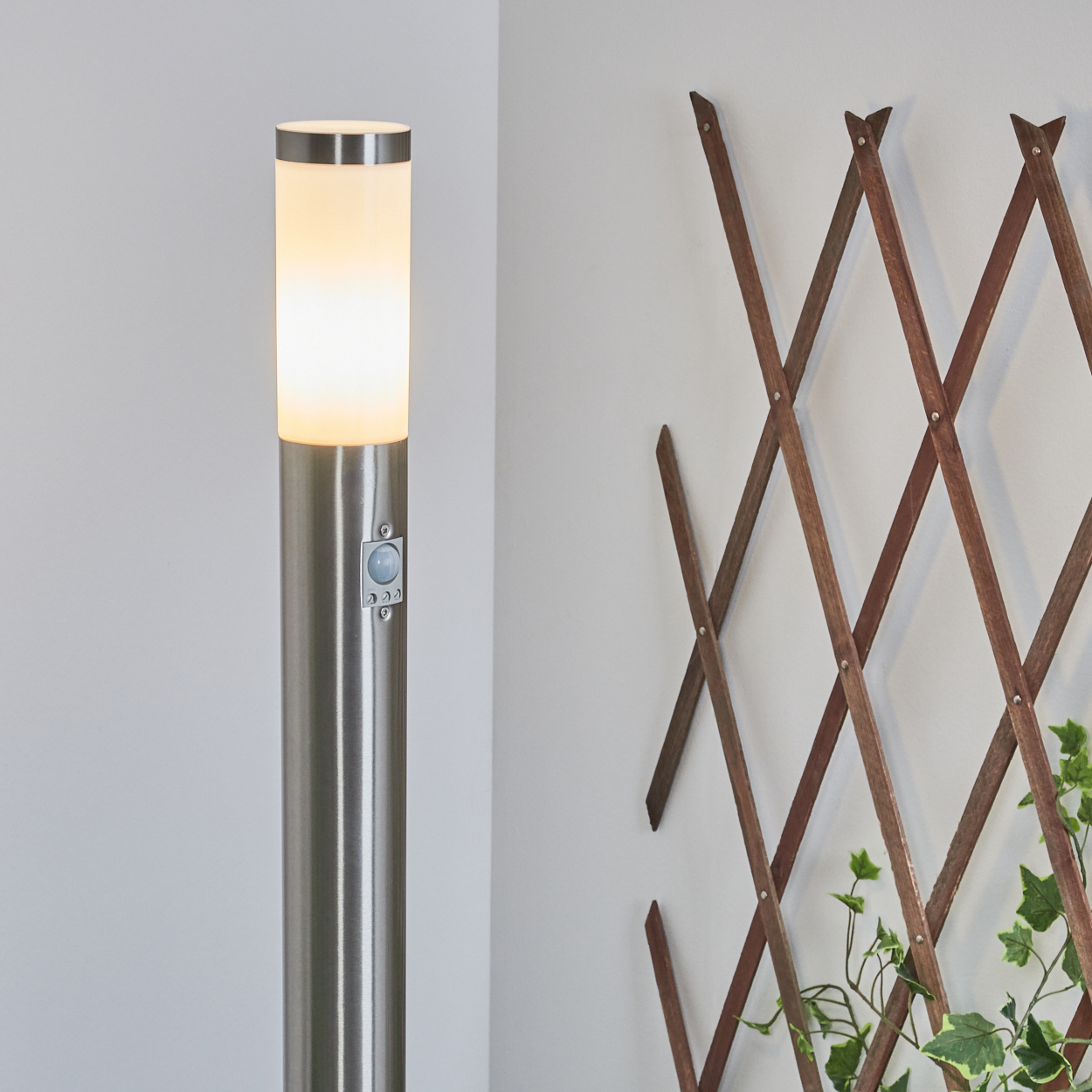 Eine Edelstahl-Außenlampe leuchtet neben einem Holzgitter mit Efeu. Die Lampe hat einen Bewegungssensor.
