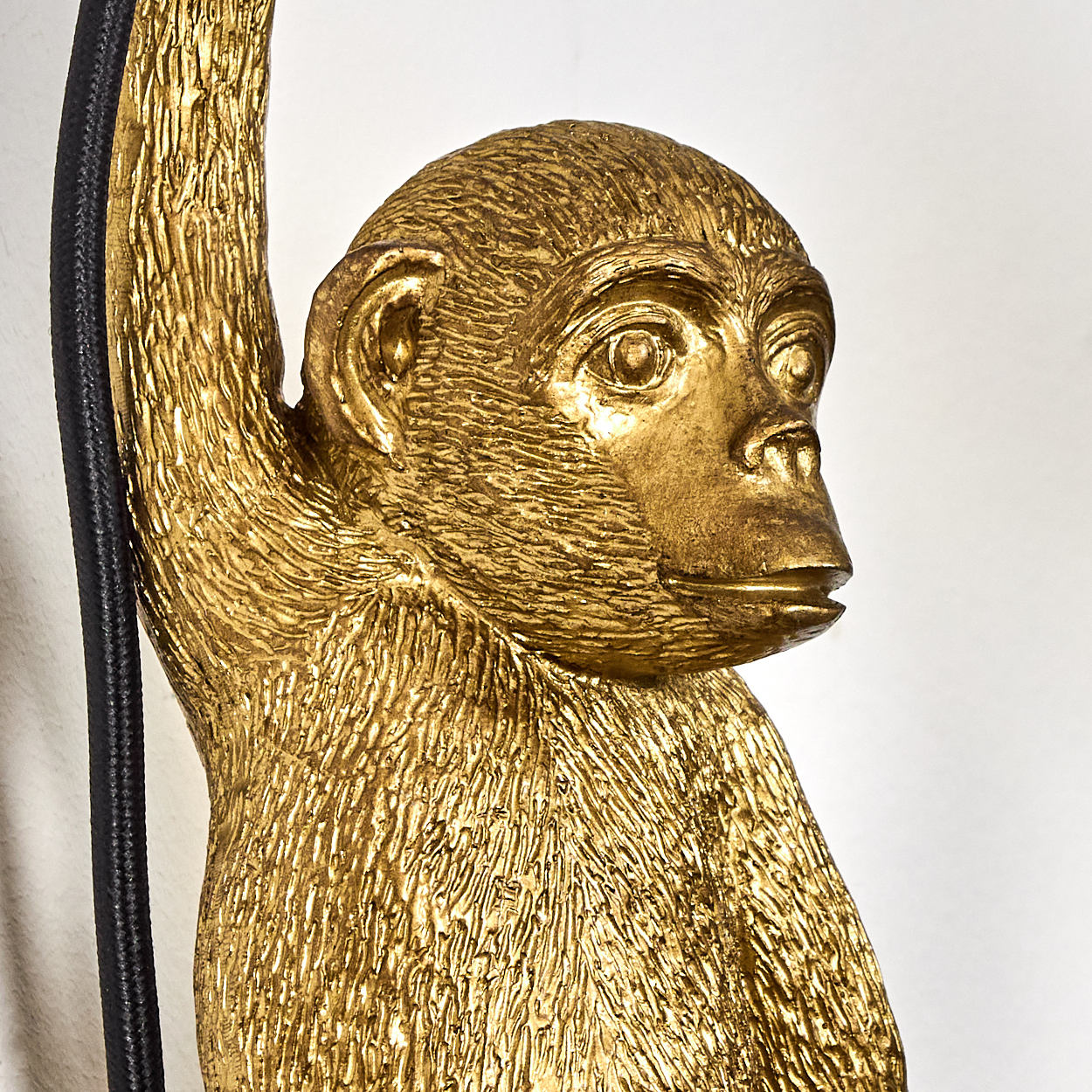 Eine goldene Affenskulptur mit detailliertem Fell, die sich an einem schwarzen Kabel festhält. Vor weißem Hintergrund blickt sie zur Seite.