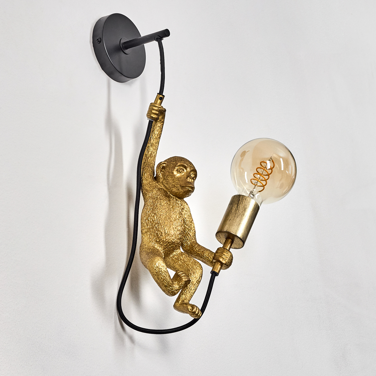 Eine goldene affenförmige Lampe hängt an einer weißen Wand. Sie hält eine Glühbirne. Ein schwarzes Kabel verbindet die Lampe mit der Wandbefestigung.