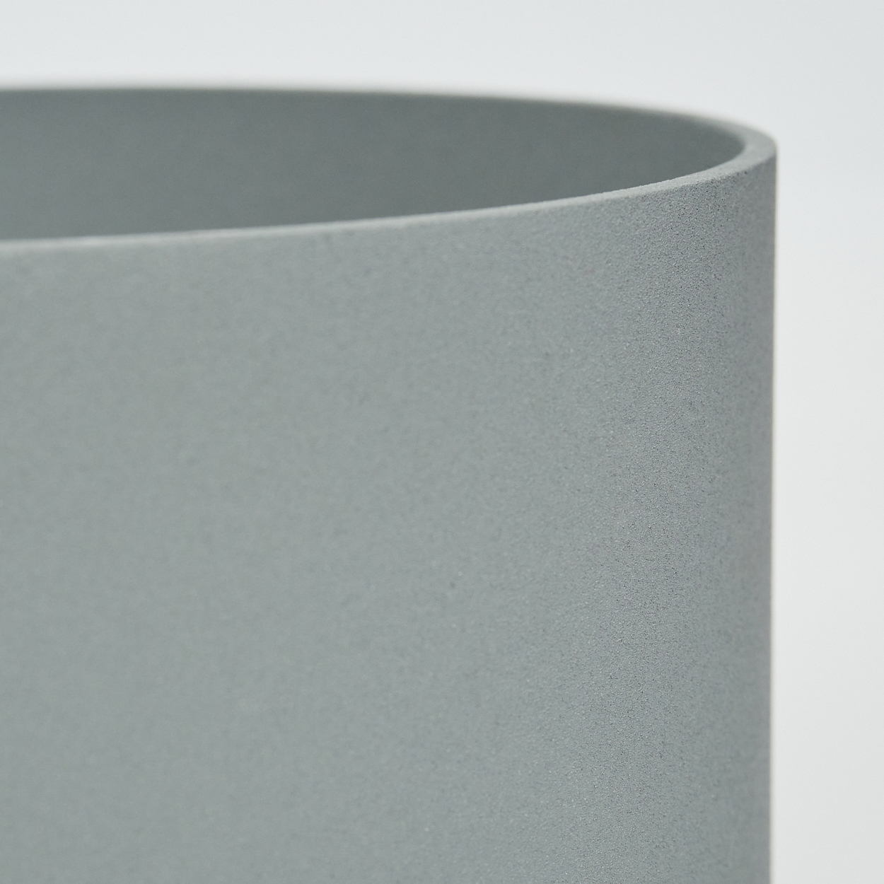Nahaufnahme eines grauen, zylindrischen Objekts, wahrscheinlich eine Vase oder ein Topf. Die Oberfläche hat eine strukturierte Optik, vor weißem Hintergrund.