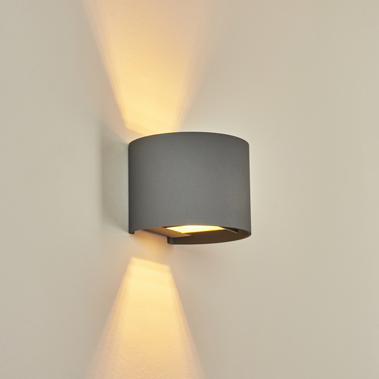 Eine graue Wandlampe ist an einer beigefarbenen Wand angebracht. Sie strahlt warmes Licht nach oben und unten aus.