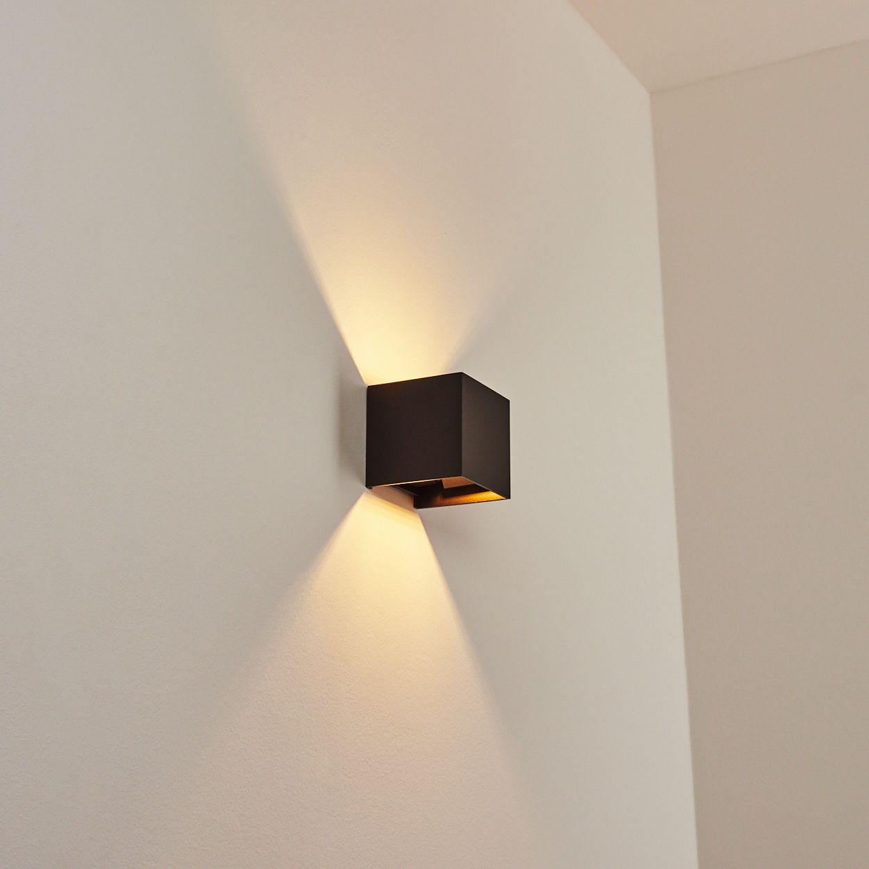 Eine schwarze, würfelförmige Wandleuchte strahlt Licht nach unten und außen auf eine hellfarbige Wand. Die Szene ist drinnen, minimalistisch.