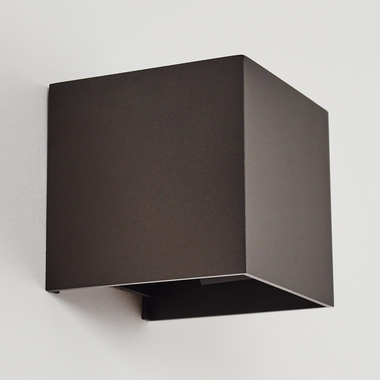 Dunkelbraune, quadratische Wandleuchte an einer weißen Wand. Die Leuchte hat eine kastenförmige Form mit offenem Boden.
