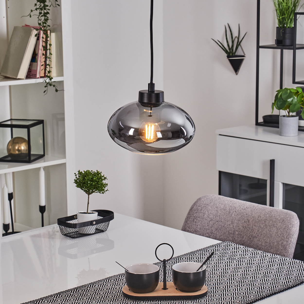 Über einem weißen Tisch hängt eine Pendelleuchte. Auf dem Tisch stehen eine kleine Pflanze, zwei Schalen und ein dekorativer Läufer.