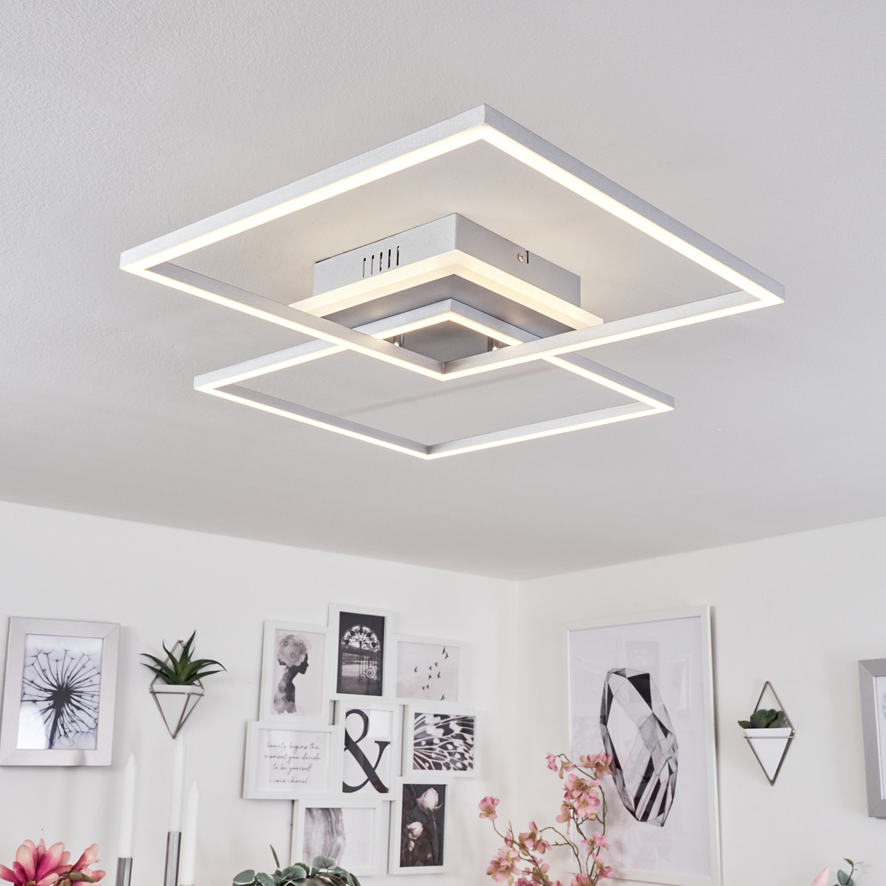Eine moderne Deckenleuchte mit geometrischem Design. Quadratische Rahmen sind beleuchtet, mit Kunst und Pflanzen an der weißen Wand darunter.