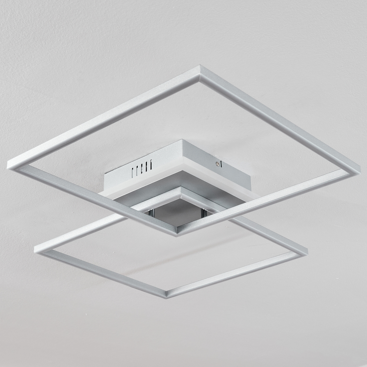 Eine moderne Deckenleuchte mit zwei sich überlappenden quadratischen Rahmen. Silbermetallkonstruktion vor weißer Decke.