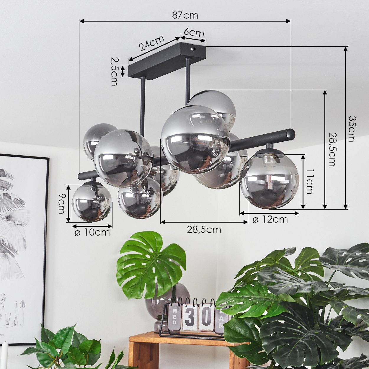 Eine moderne Deckenleuchte mit mehreren kugelförmigen Glasschirmen, ein Holzregal mit Pflanzen, ein Kalender und ein gerahmtes Kunstwerk an der Wand.