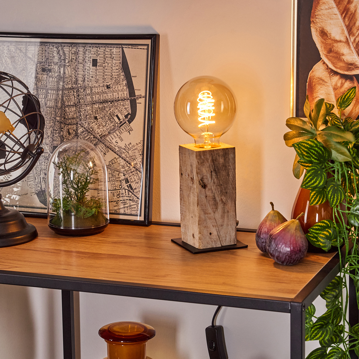 Ein Holztisch mit Lampe, Karte, Feigen und Pflanzen. Die Lampe hat eine große Glühbirne. Eine Glaskuppel mit Pflanzen steht auf dem Tisch.