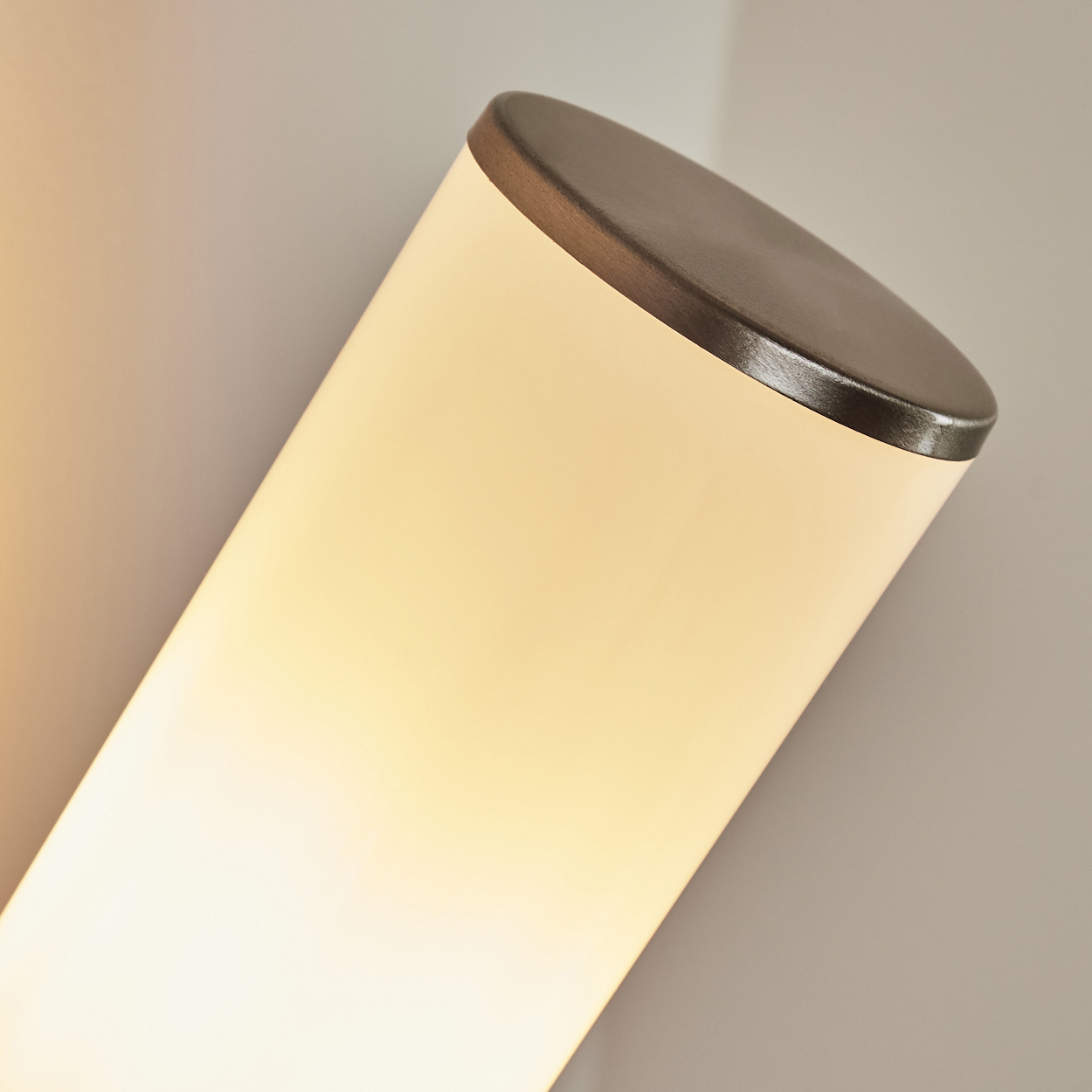 Eine zylindrische Lampe ist schräg im Bild. Sie ist cremefarben mit einem dunklen Metallaufsatz. Der Hintergrund ist hell neutral.