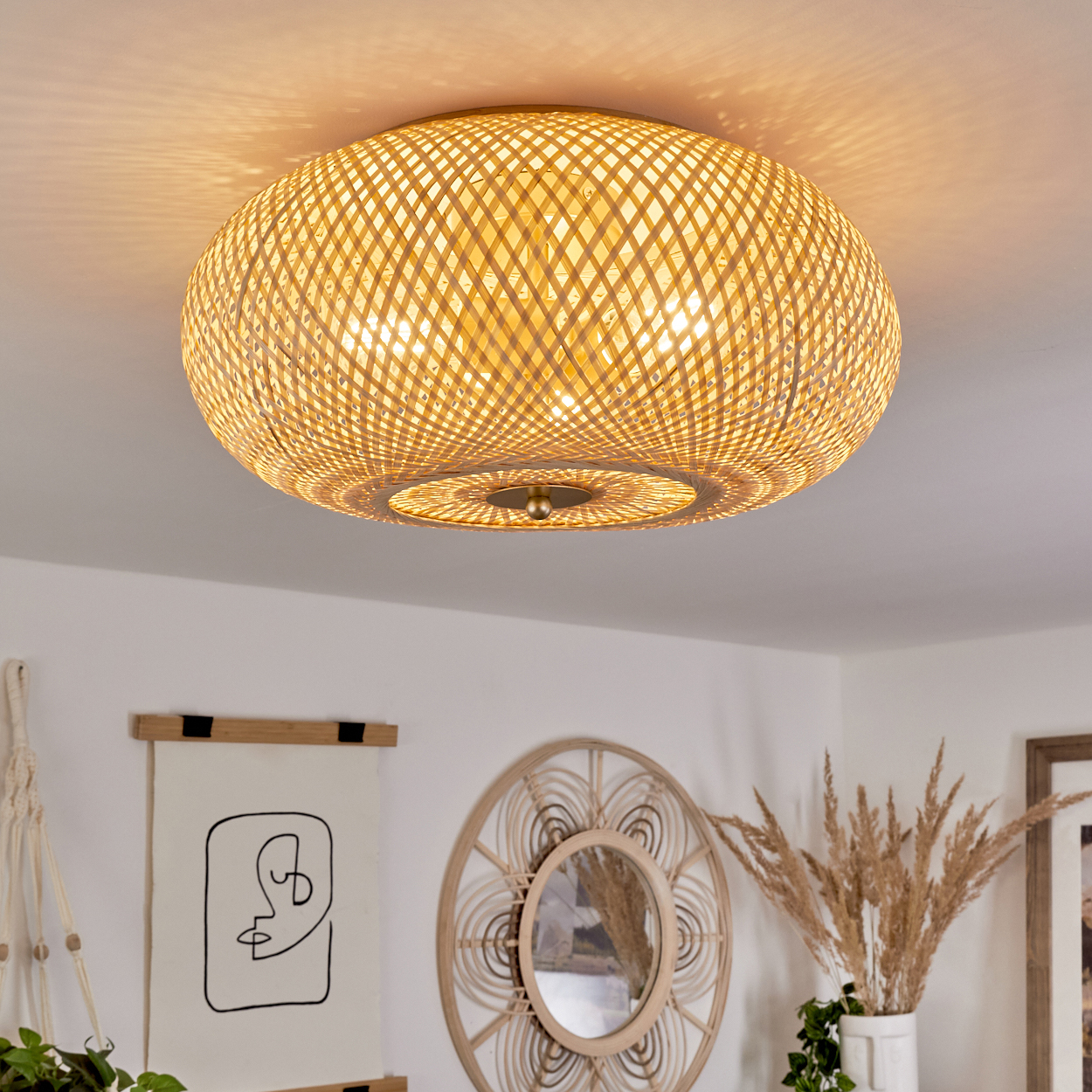 Eine gewebte Deckenlampe aus Rattan beleuchtet einen Raum. Darunter dekorieren ein Spiegel, ein Kunstdruck und Pflanzen den Raum. Warmes Licht füllt die Szene.