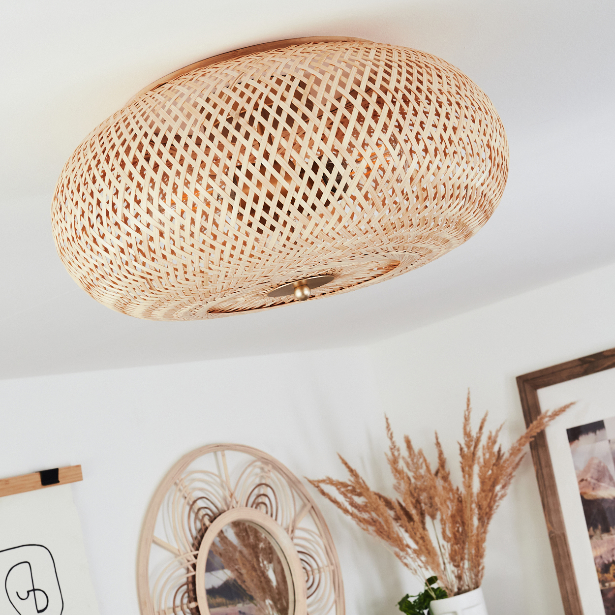 Gewebte Rattan-Lichtvorrichtung an einer weißen Decke. Dekorative Spiegel, gerahmte Kunst und getrocknete Pflanzen darunter.