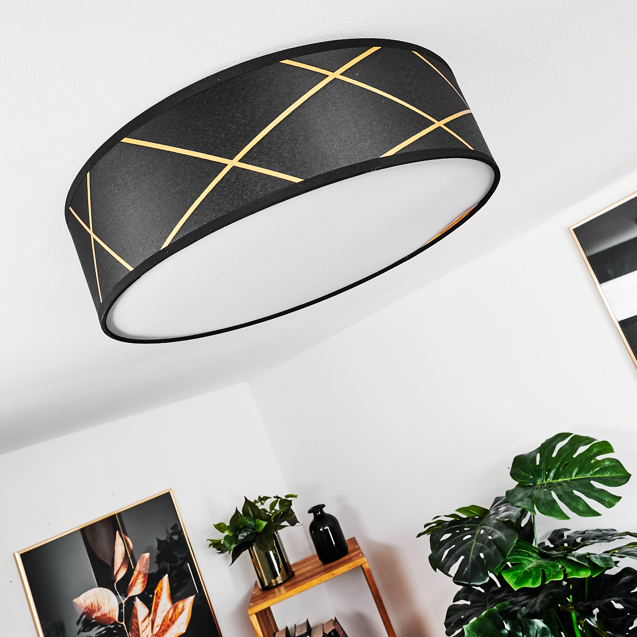 Schwarze und goldene Deckenlampe unter einer weißen Decke. Grüne Pflanzen und Holzregal mit Vasen und einem Bilderrahmen.