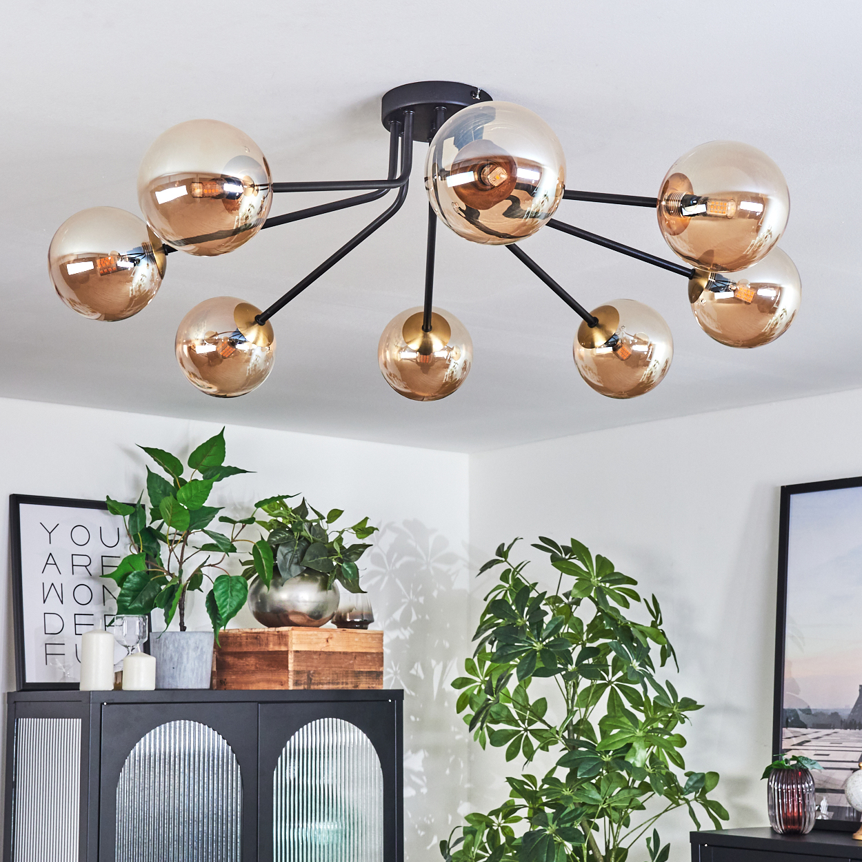 Eine moderne Deckenleuchte mit goldenen Kugeln und schwarzen Armen. Pflanzen und Möbel befinden sich in einem Raum. Ein gerahmtes Kunstwerk ist an der Wand.