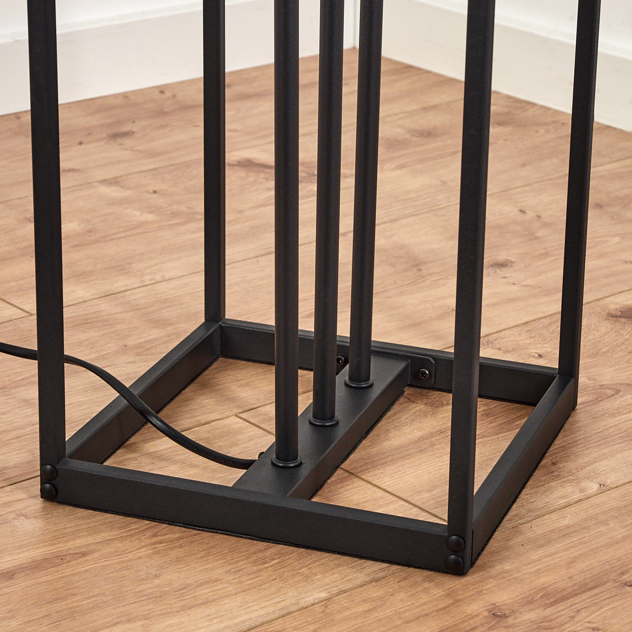 Ein schwarzer Metall-Lampensockel mit vertikalen Stäben. Der Sockel ist quadratisch, mit einem Netzkabel. Der Boden ist holzgetäfelt.