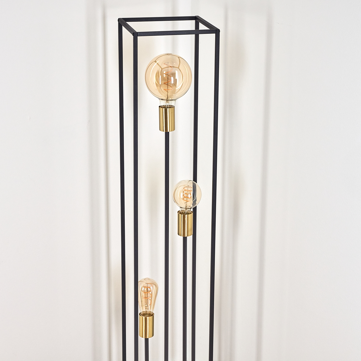 Eine schwarze Metalllampe mit drei goldfarbenen Glühbirnen vor einer weißen Wand.