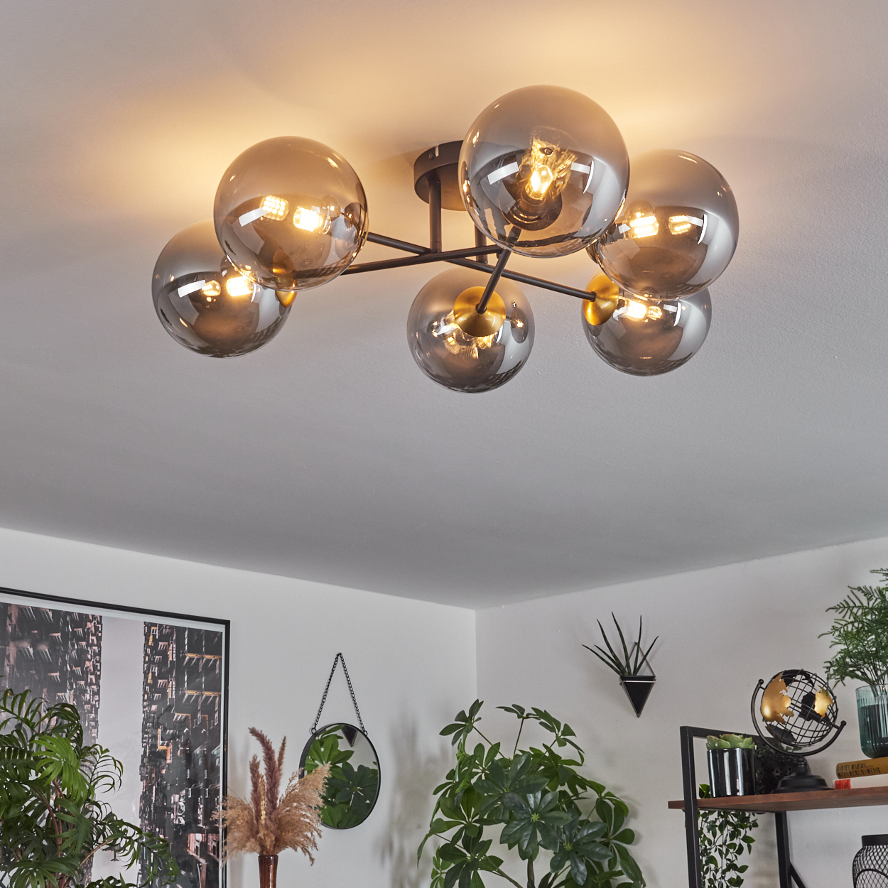 Eine Deckenleuchte mit sechs grauen Glaskugeln leuchtet. Die Szene umfasst Pflanzen, einen Spiegel und ein gerahmtes Bild, alles in einem Wohnzimmer.