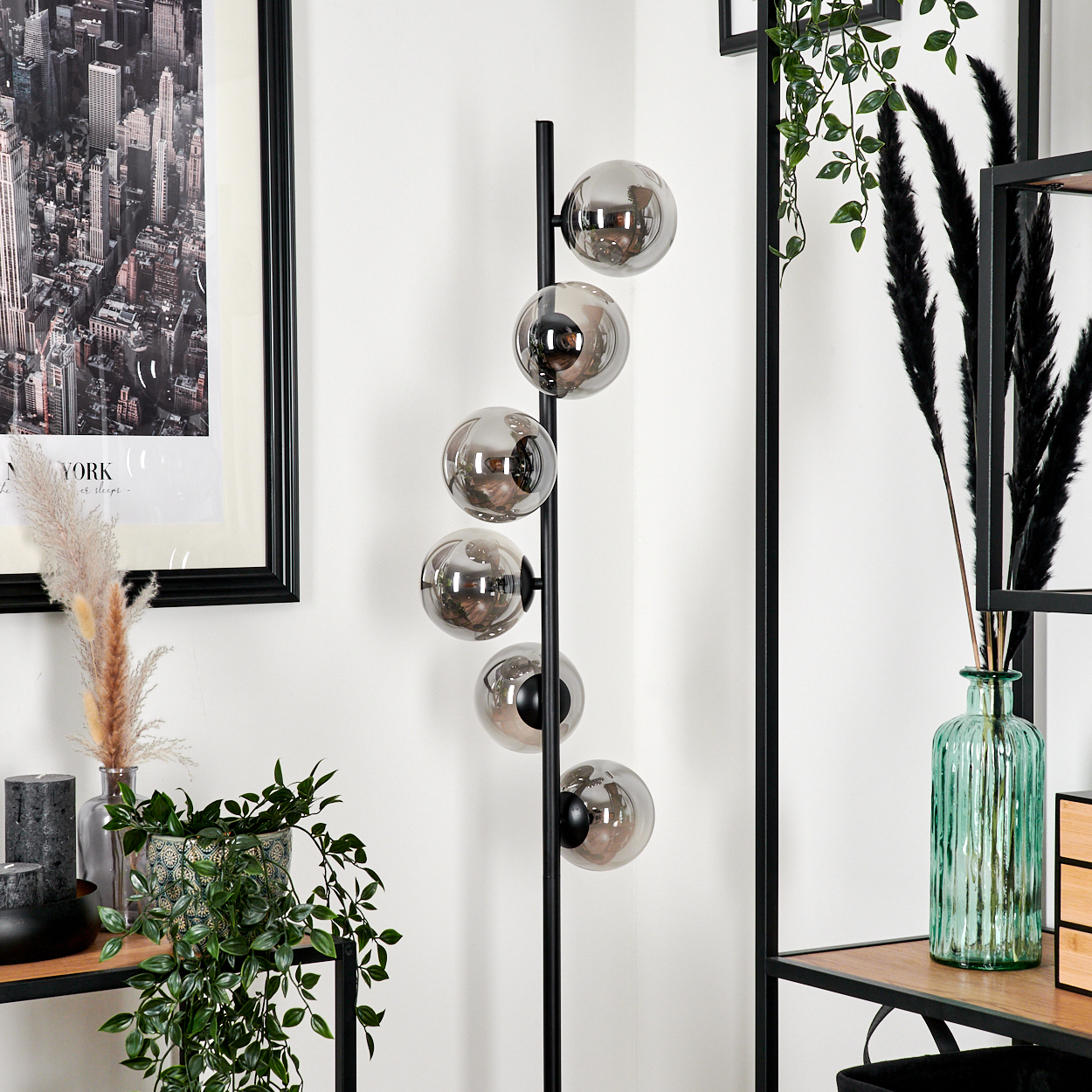 Ein Raum mit einem schwarz gerahmten Bild von New York, einer schwarzen Stehlampe mit Glaskugeln, Pflanzen und Regalen.
