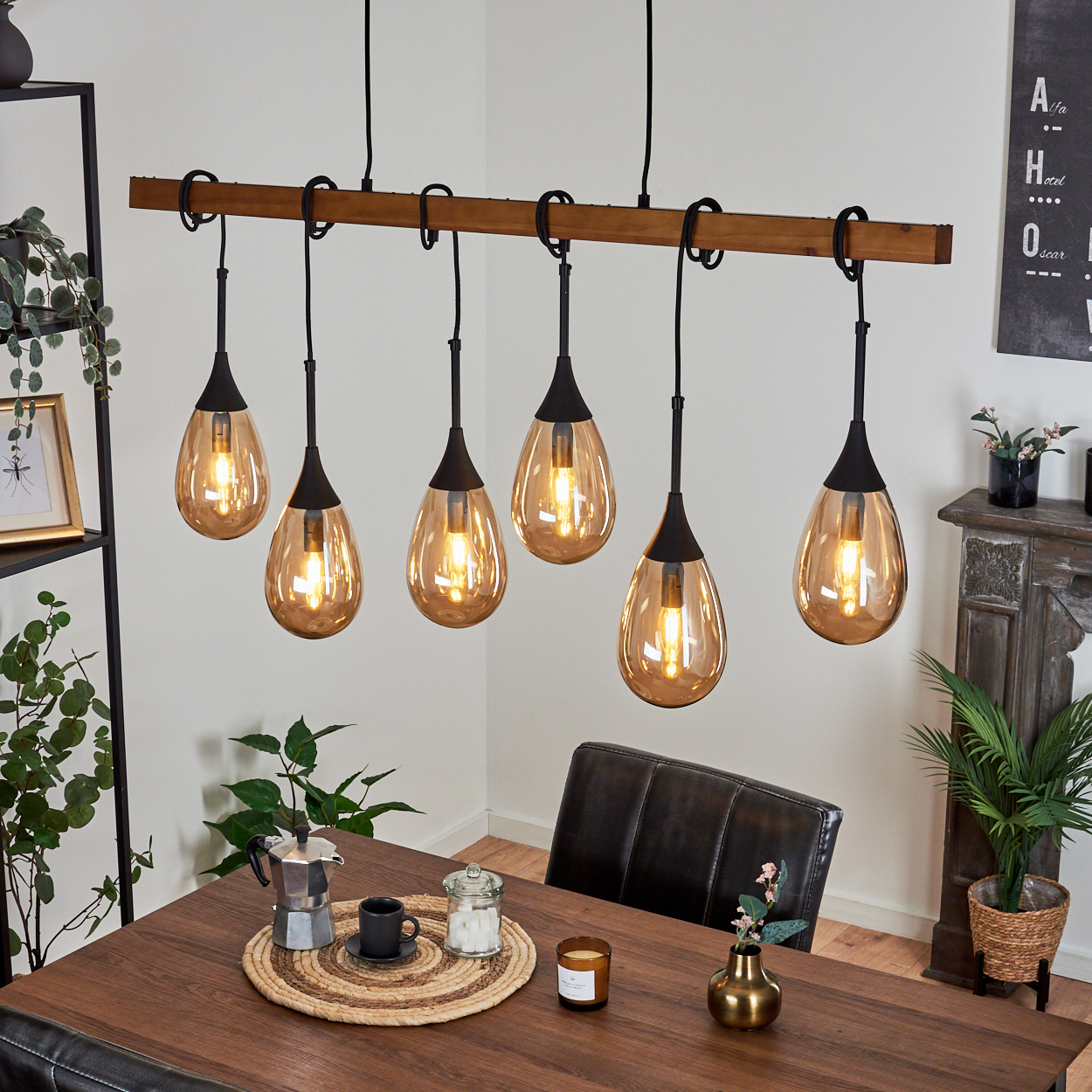 Ein Esszimmer mit Holztisch und Stühlen. Sechs Hängelampen hängen darüber. Es gibt Pflanzen, eine Kaffeemaschine und eine Kerze.