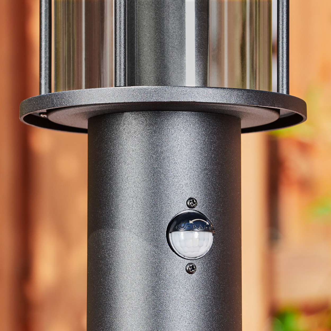 Nahaufnahme einer schwarzen Laterne mit Sensor. Der obere Teil hat einen silbernen Zylinder. Der Hintergrund ist verschwommen, eine braune Wand.