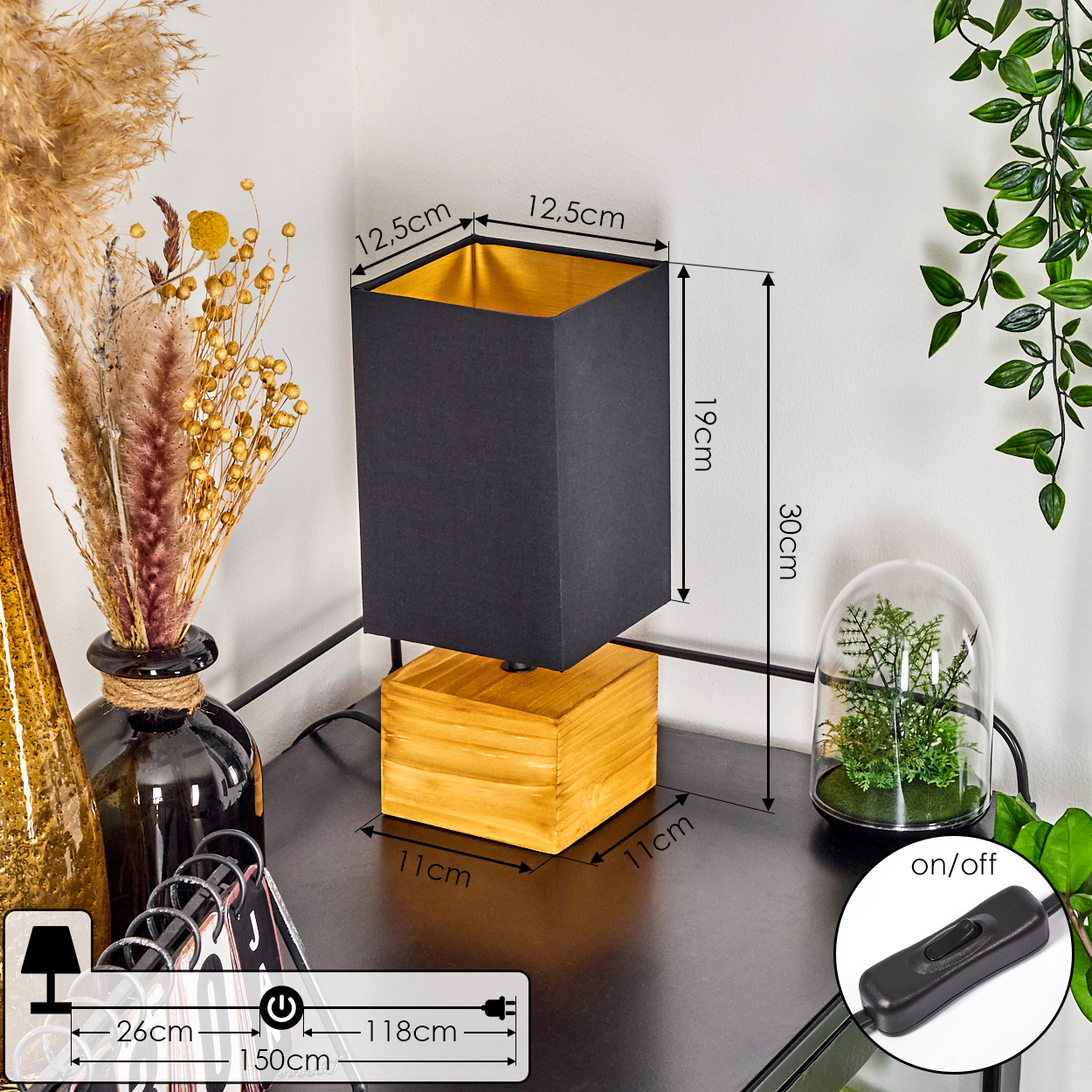 Eine schwarze Lampe mit Holzsockel steht auf einer dunklen Oberfläche. Maße und Details sind beschriftet. Trockenpflanzen und ein kleines Terrarium sind ebenfalls vorhanden.