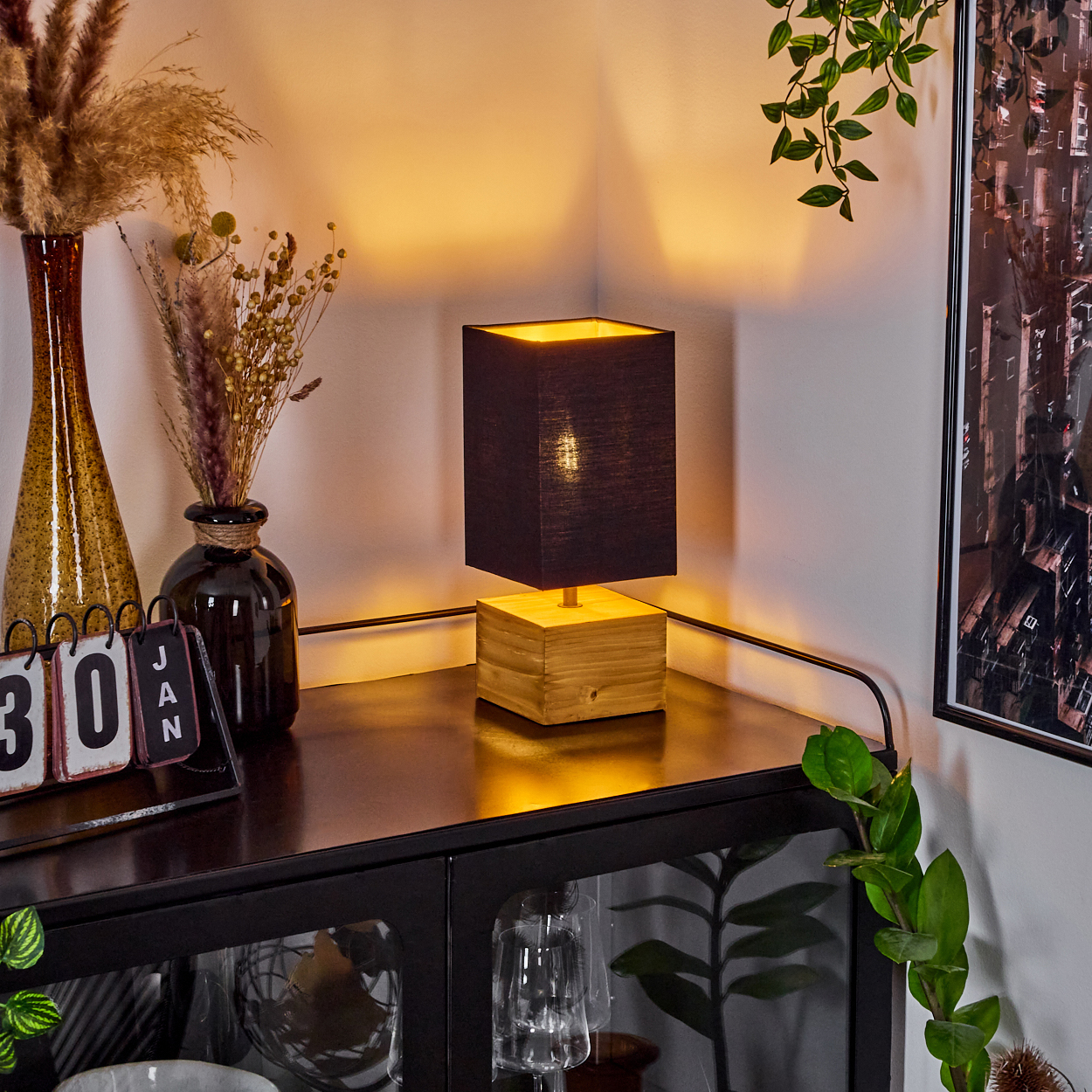 Eine Lampe mit schwarzem Schirm und Holzsockel steht auf einem dunklen Schrank. Vasen und ein Kalender stehen auf dem Schrank. Pflanzen hängen darüber.