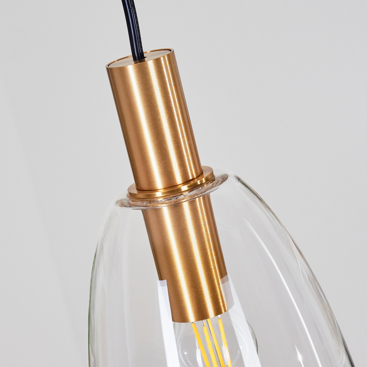 Nahaufnahme einer modernen Pendelleuchte. Goldfarbene zylindrische Halterung mit klarem Glasschirm. Ein schwarzes Kabel hängt von oben.