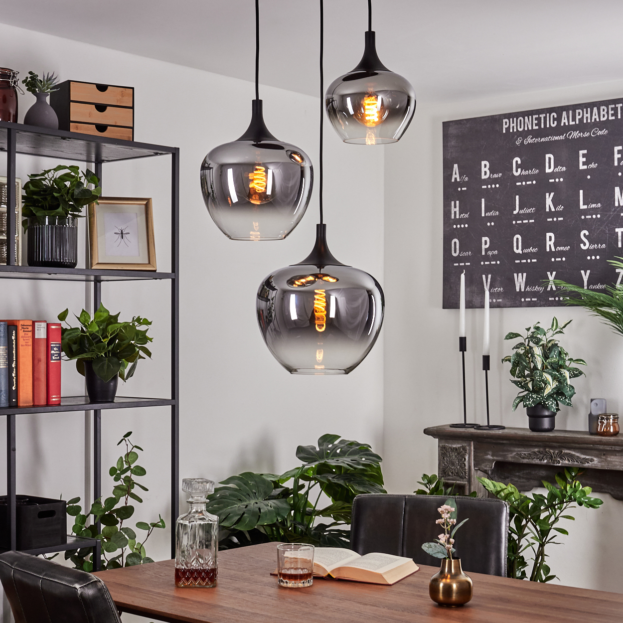 Drei Hängelampen hängen über einem Esstisch. Pflanzen, eine Karaffe und Bücher sind sichtbar. Eine phonetische Alphabet-Tafel hängt an der Wand.