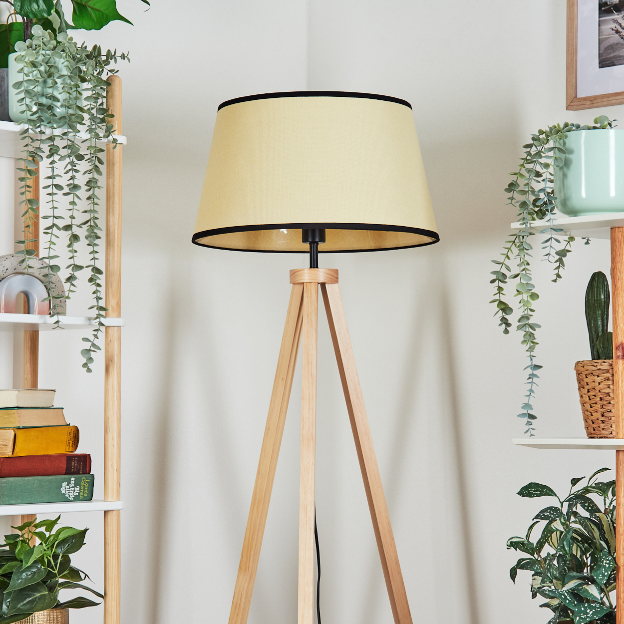 Eine Stehlampe aus Holz mit beigem Schirm, umgeben von Pflanzen auf Regalen. Die Wand ist weiß.