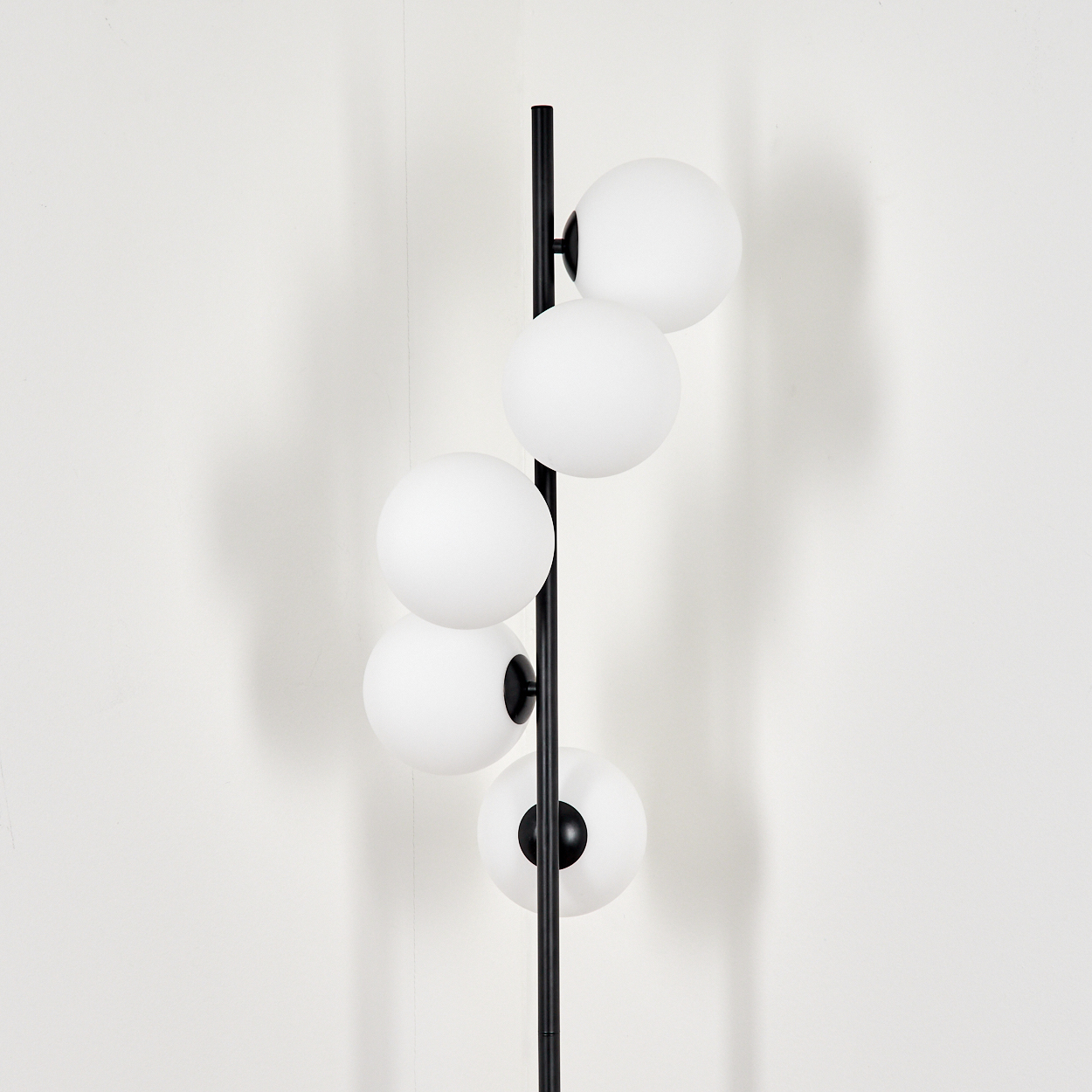 Eine Stehlampe mit schwarzem Metallmast und fünf kugelförmigen weißen Glasschirmen. Der Hintergrund ist eine schlichte weiße Wand.