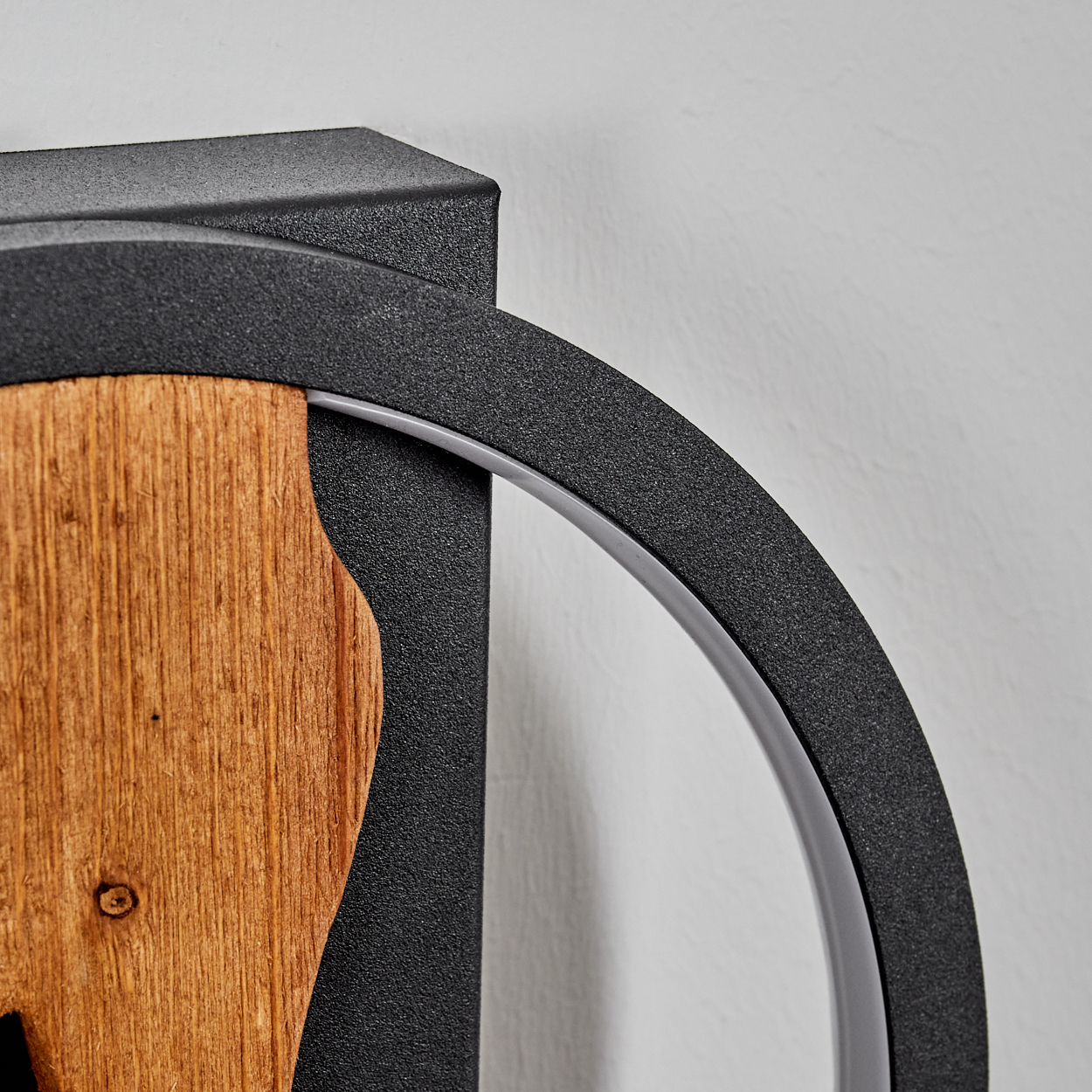Nahaufnahme eines dekorativen Stücks mit Holz und schwarzem Metall. Ein gebogener Metallrahmen umgibt das Holzelement vor einer weißen Wand.