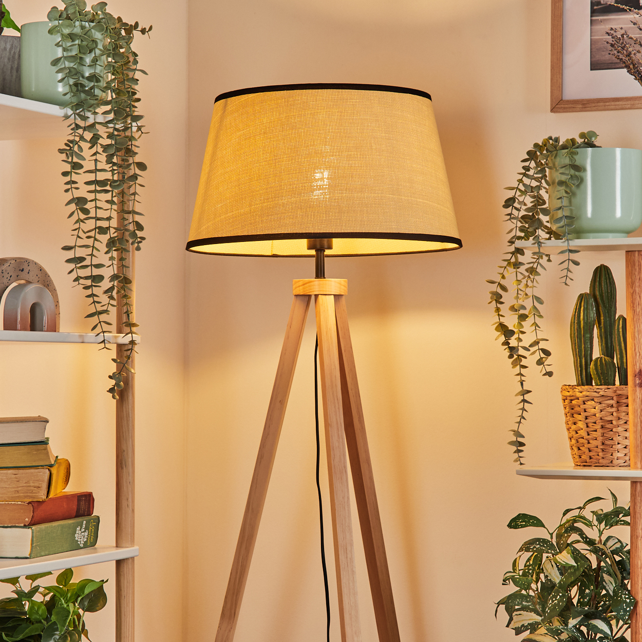 Holz-Stativlampe mit beigem Schirm. Regale mit Pflanzen, Büchern und einem Kaktus sind sichtbar. Weiche, warme Beleuchtung.