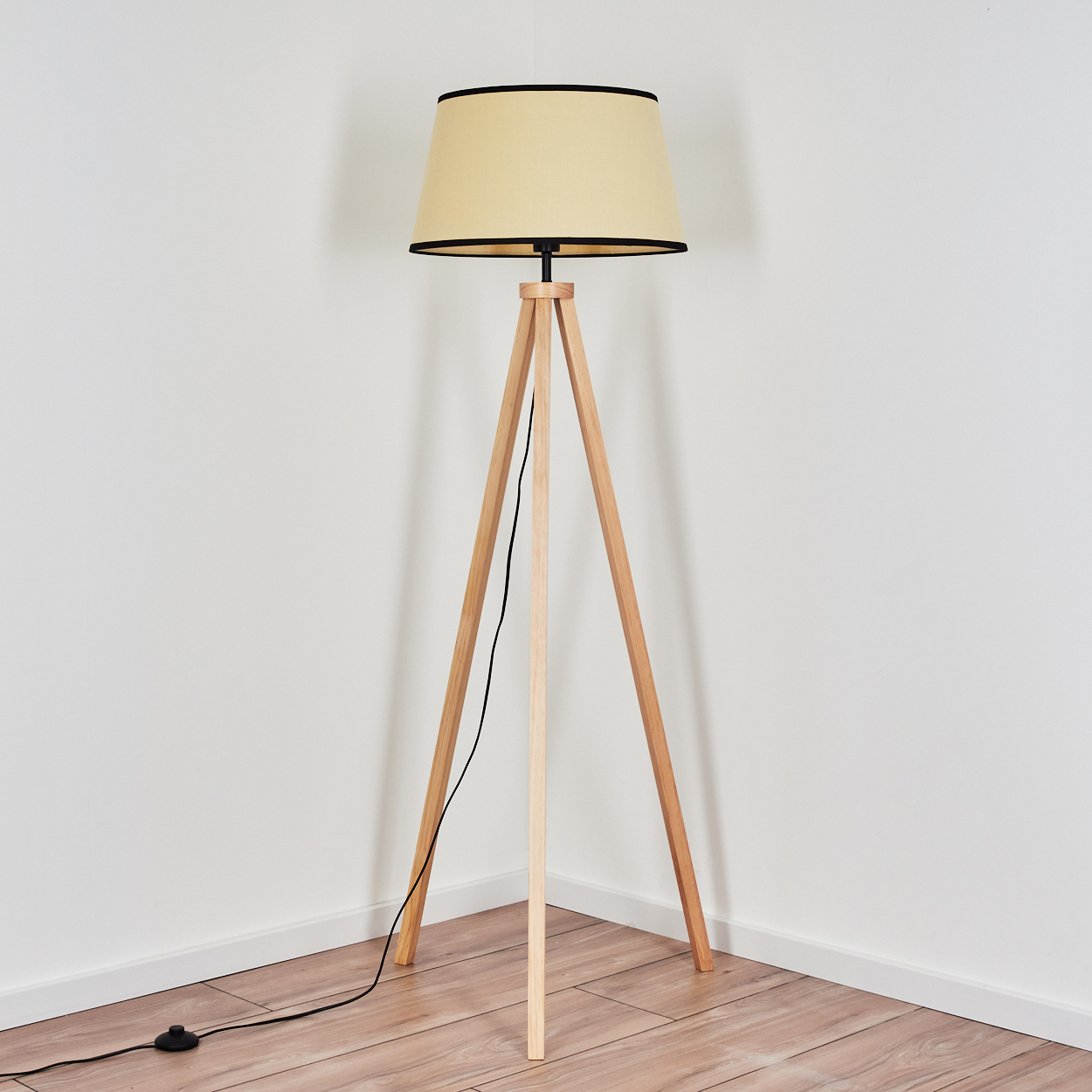 Eine Holz-Stativlampe mit beigem Lampenschirm und schwarzem Rand steht in einer Ecke. Der Boden ist aus Holz, die Wände sind weiß.