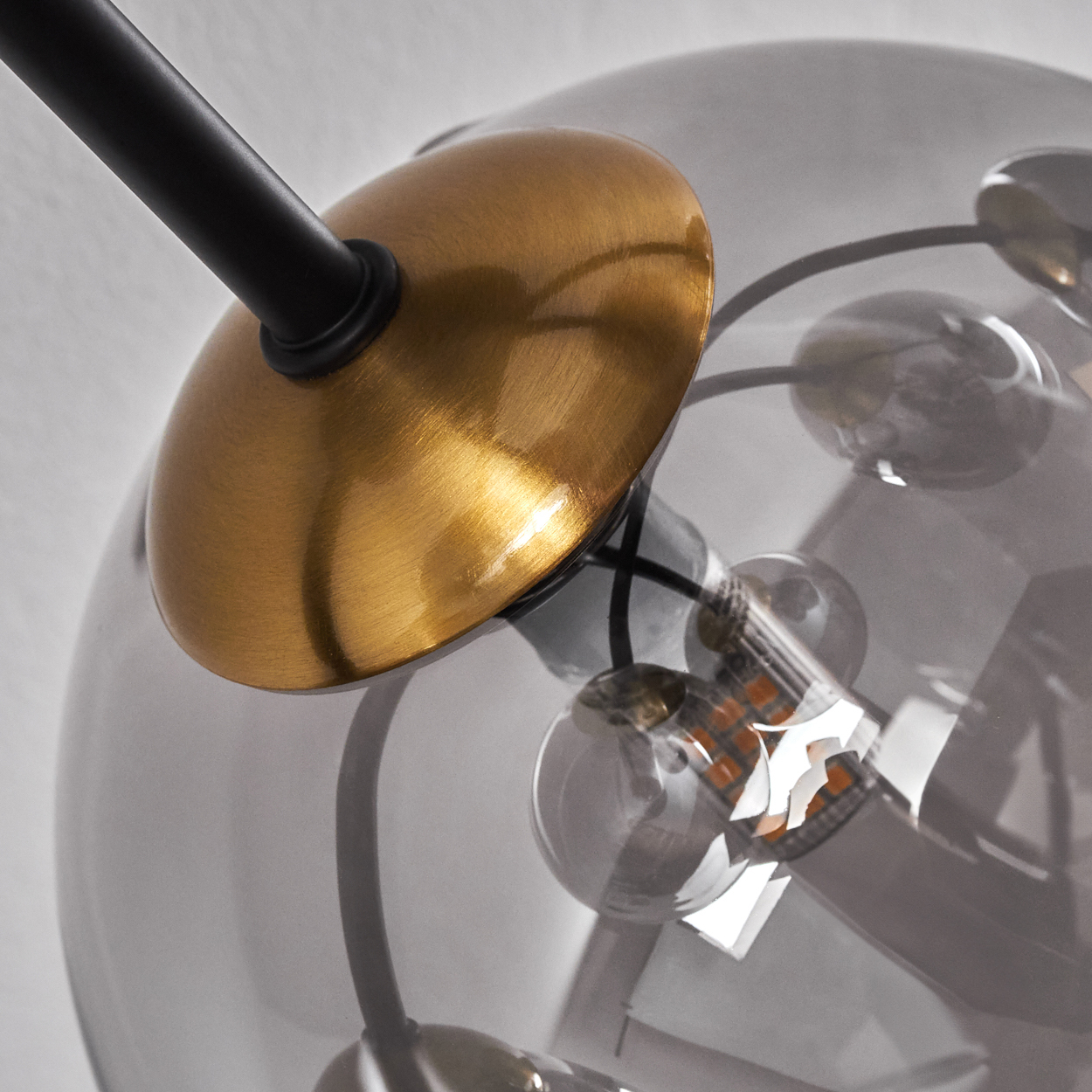 Nahaufnahme eines modernen Kronleuchters. Er hat eine goldfarbene Metallscheibe, schwarze Stäbe und transparente, runde Glaskugeln. Der Hintergrund ist weiß.