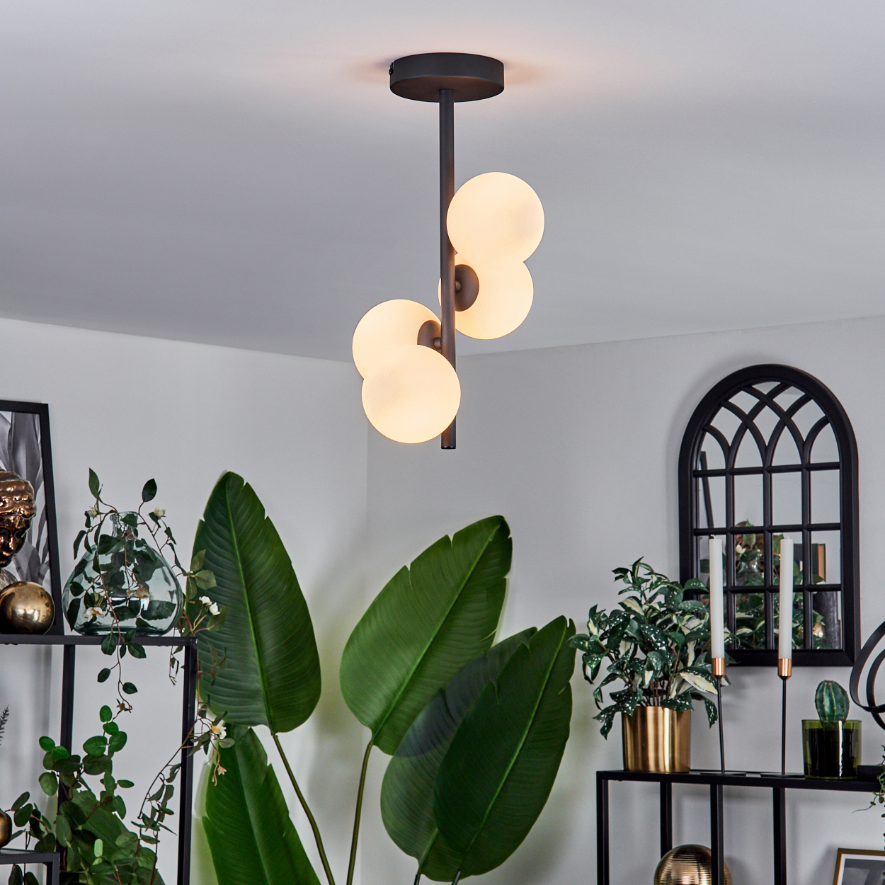 Ein modernes Wohnzimmer mit einer Hängeleuchte. Große grüne Pflanzen, ein schwarzes Regal und ein dekoratives Fenster sind ebenfalls vorhanden.