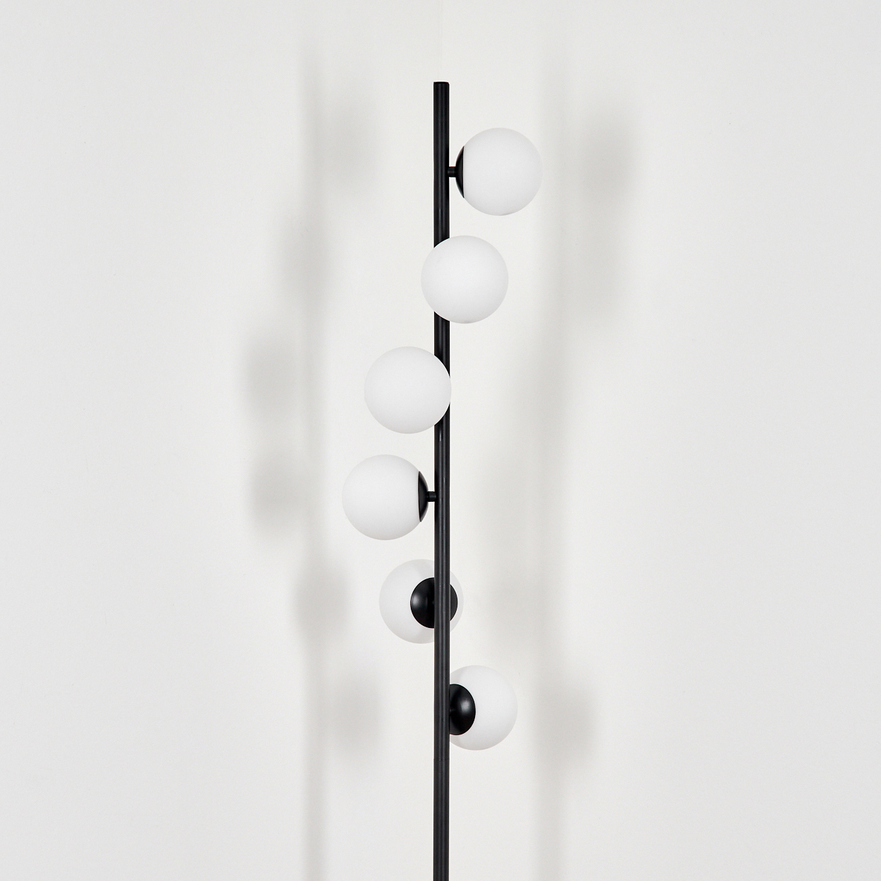 Eine schwarze Stehlampe mit sechs weißen, kugelförmigen Lampenschirmen. Die Lampe steht vor einer weißen Wand, auf der ein Schatten geworfen wird.