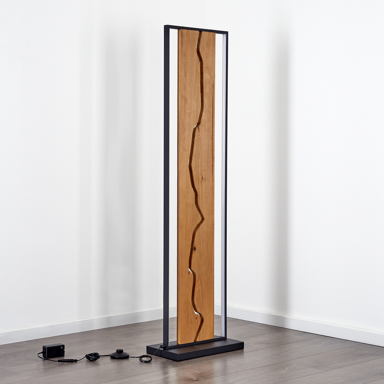 Eine hohe Holz-Metall-Lampe steht vor einer weißen Wand. Die Holzmitte hat ein geschnitztes Design. Schwarzer Sockel und Rahmen.