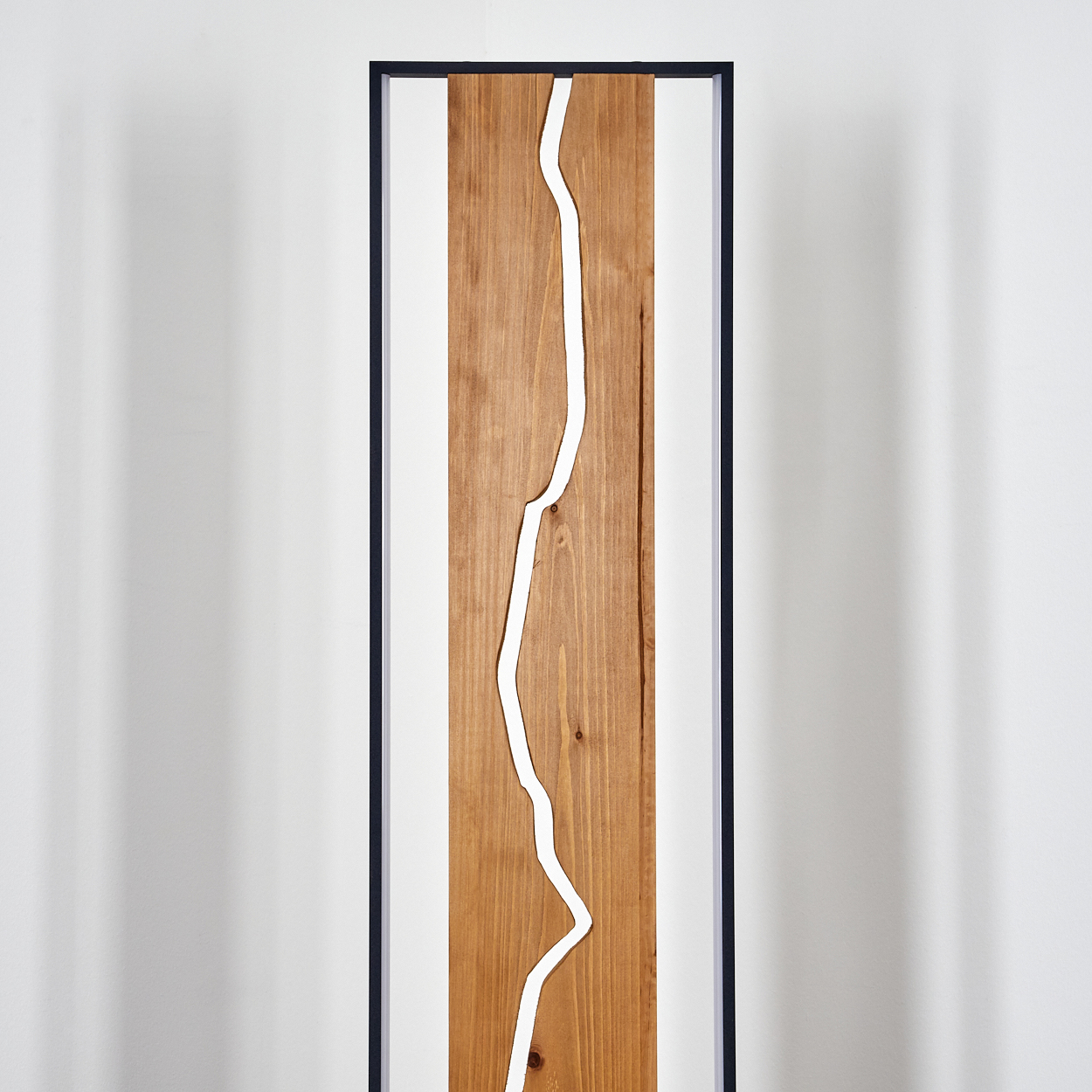 Holzkunstwerk mit einer weißen, wellenförmigen Linie in der Mitte, schwarz umrahmt. Vor einer weißen Wand, von oben beleuchtet.